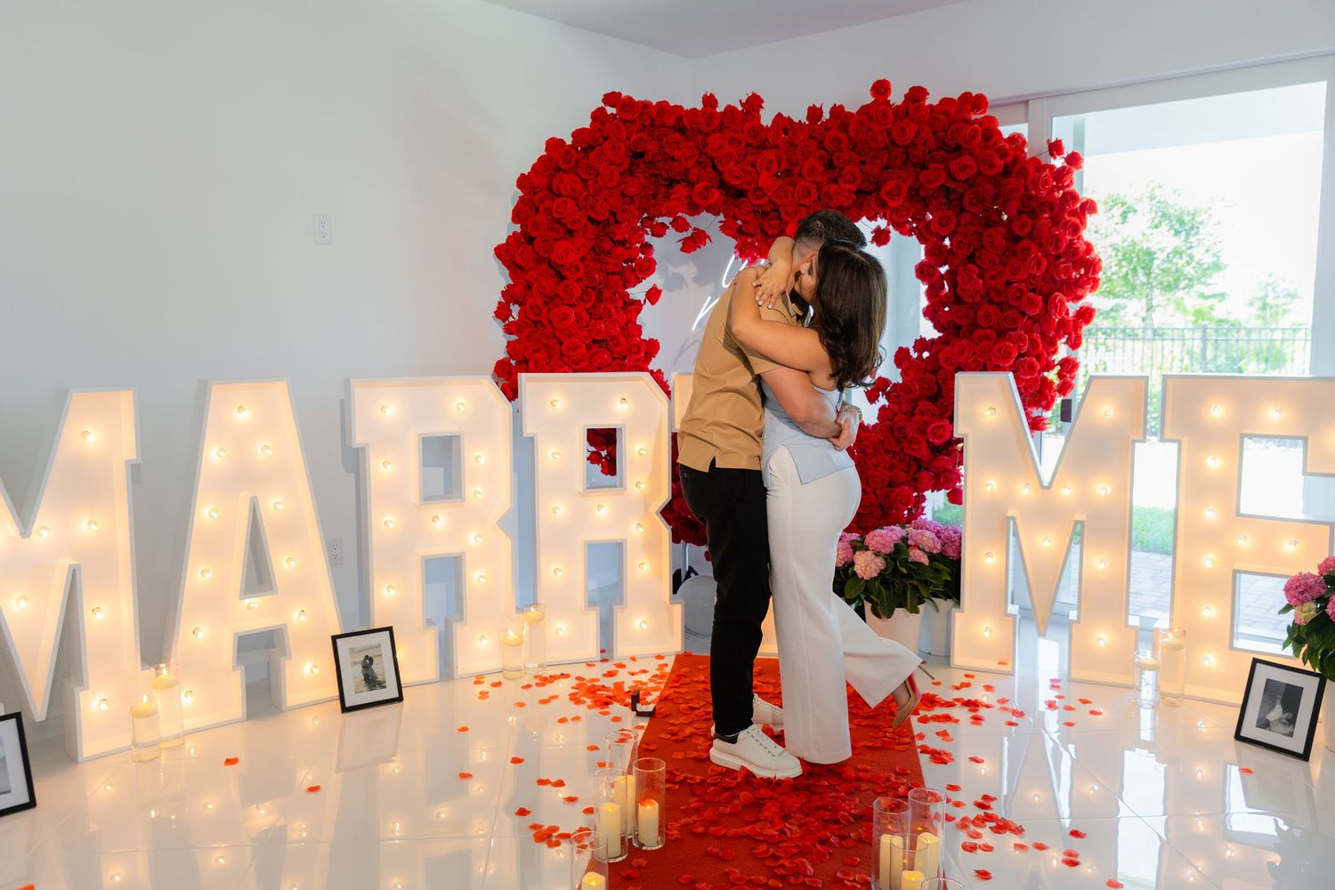 Glowing MARRY ME letters surrounded by white candles and red rose petals create an intimate proposal scene.