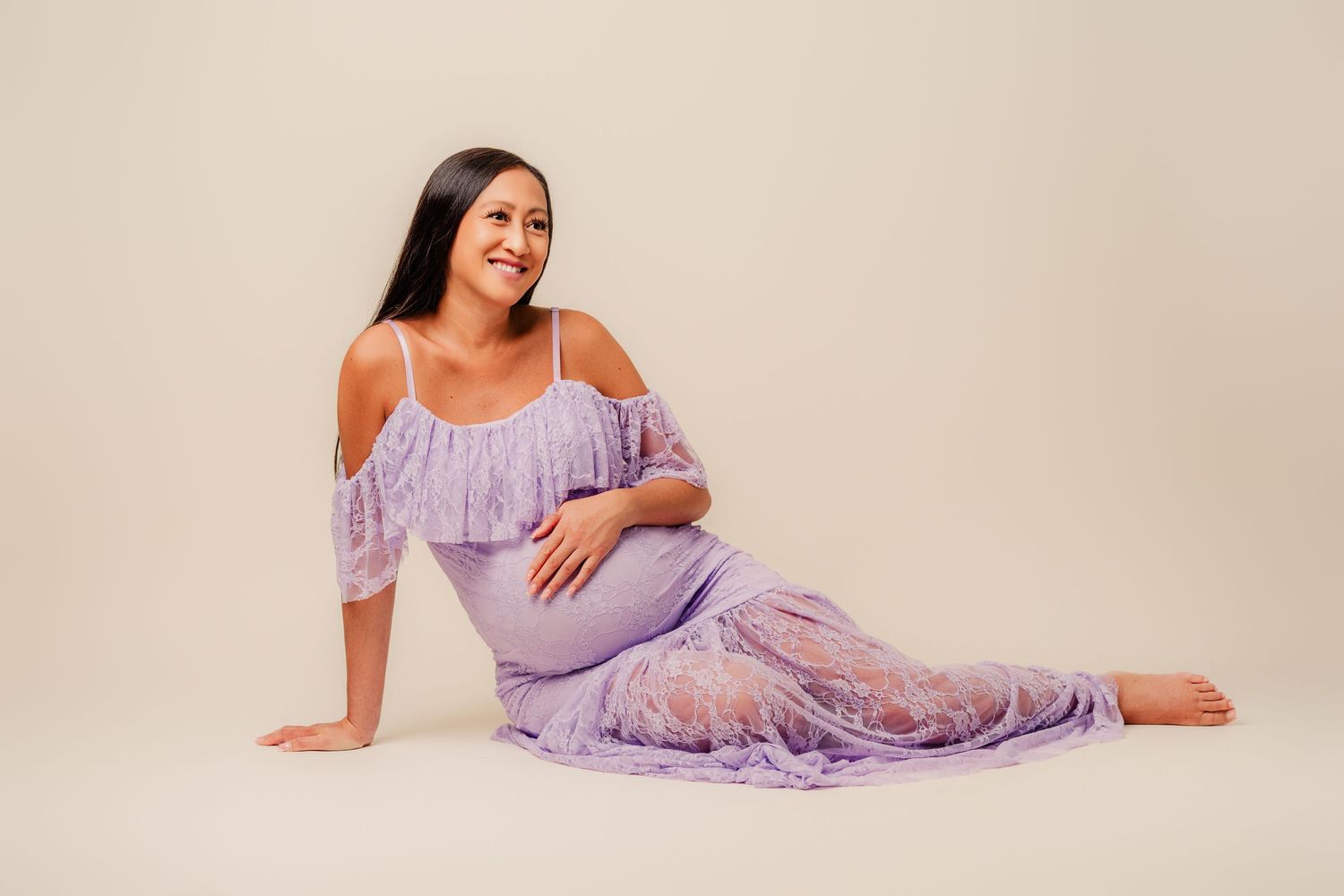 Woman in lavender lace dress poses gracefully on beige backdrop in elegant photoshoot series.
