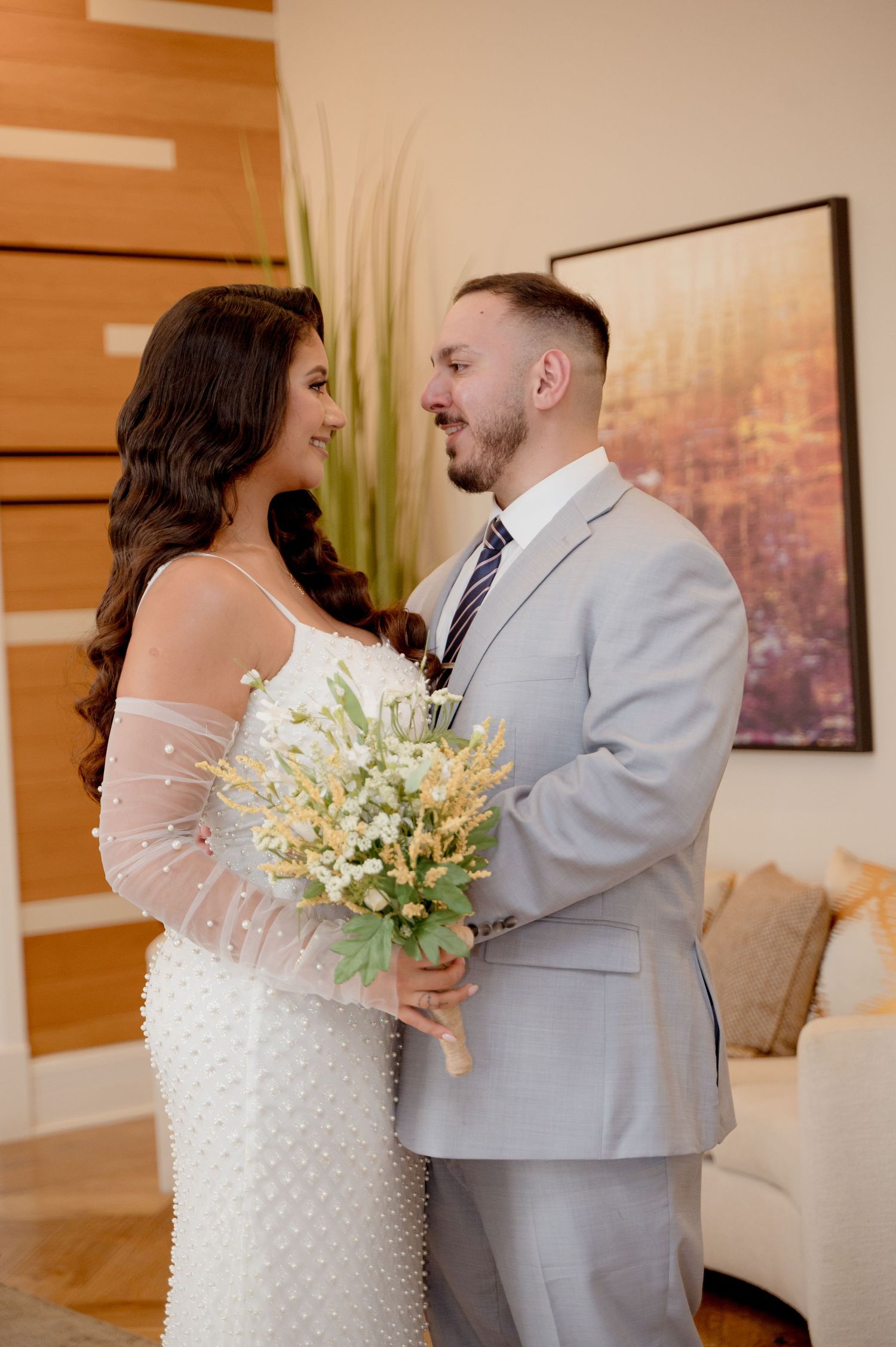 A couple shares a tender moment in a sunlit room, with the bride wearing a textured white gown and holding a floral bouquet.