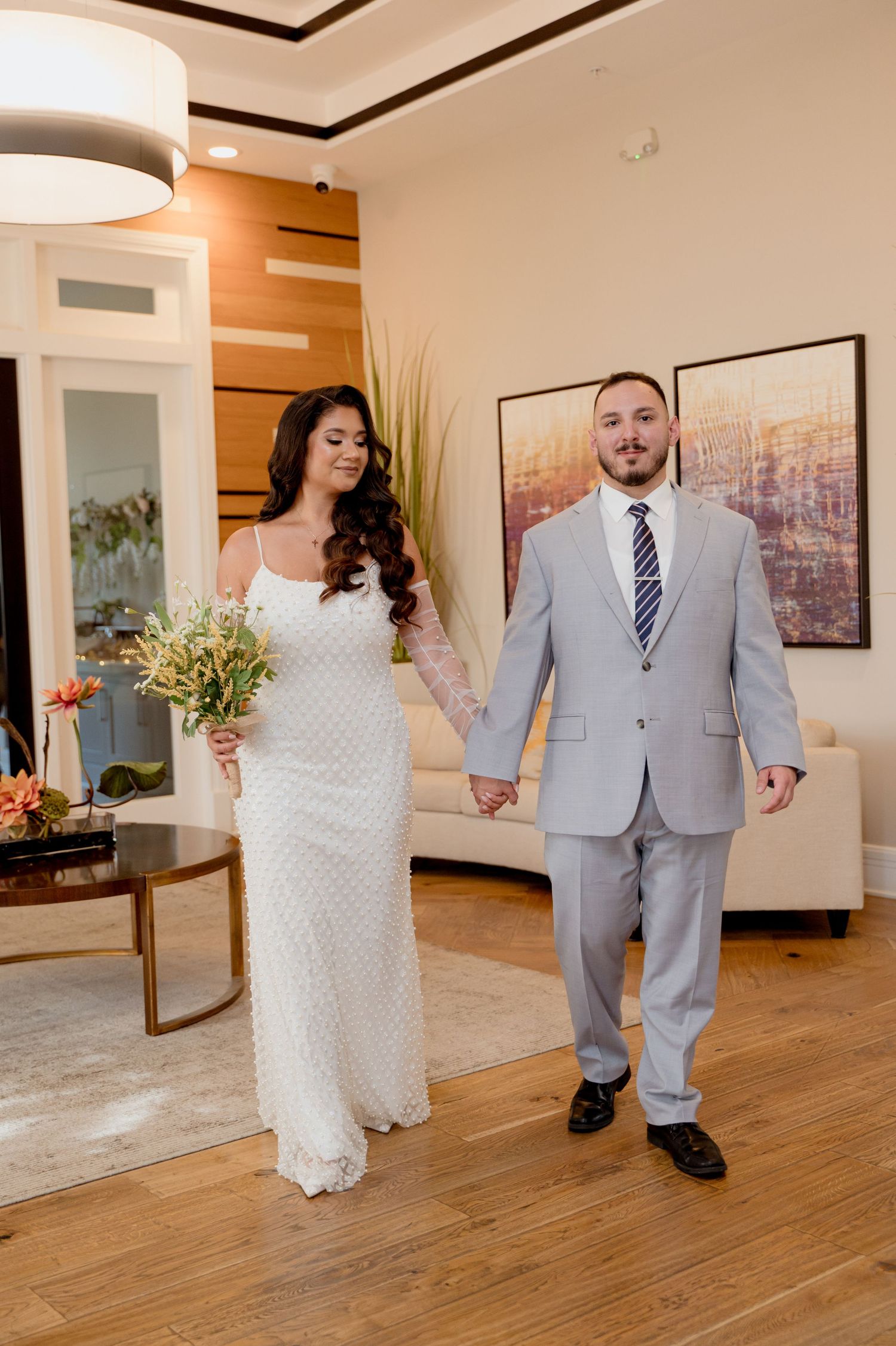 A couple in wedding attire dances together on a hardwood floor in a bright indoor venue.