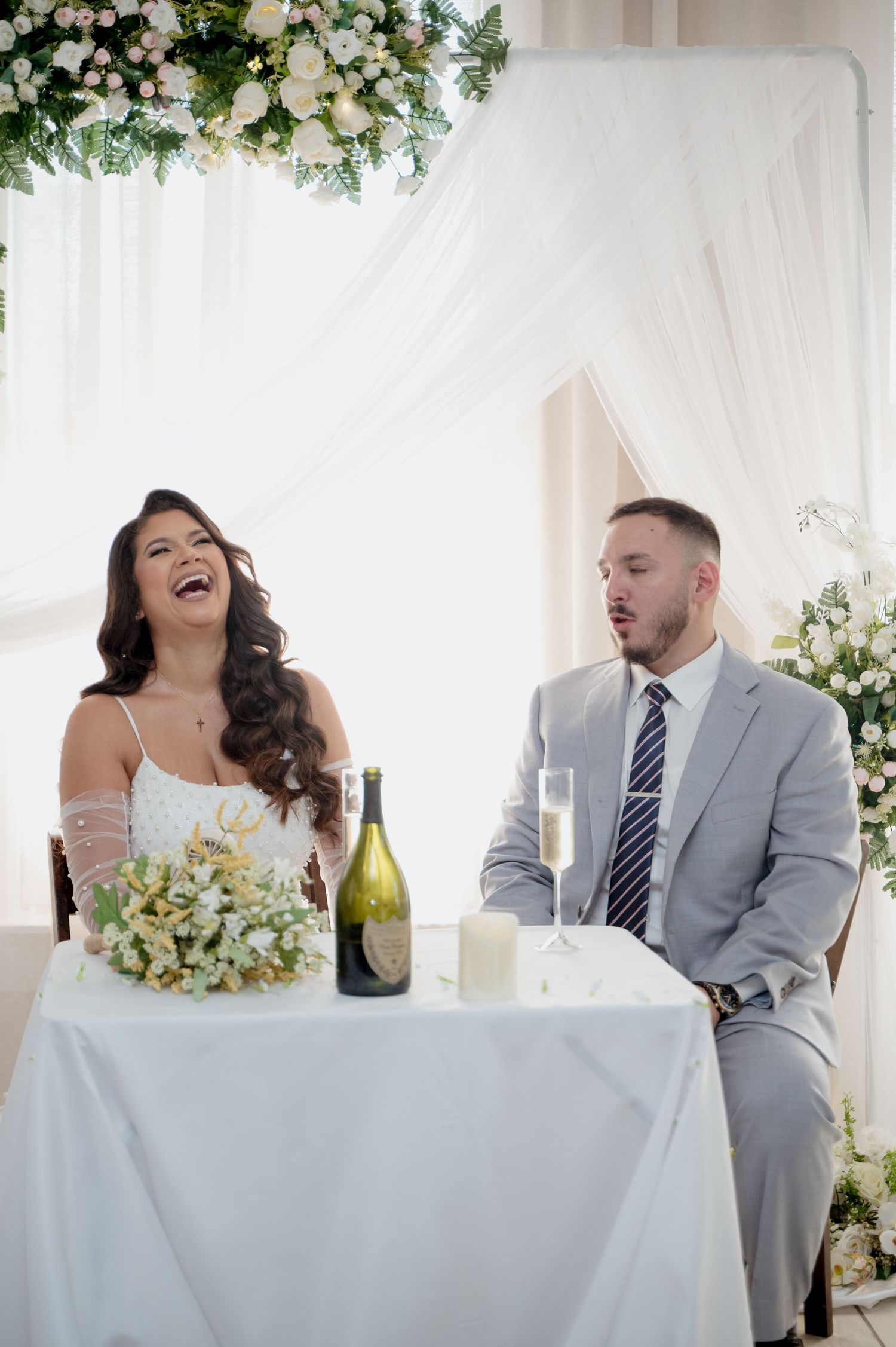 A joyful wedding moment captured at a white-decorated reception table with floral arrangements and champagne.
