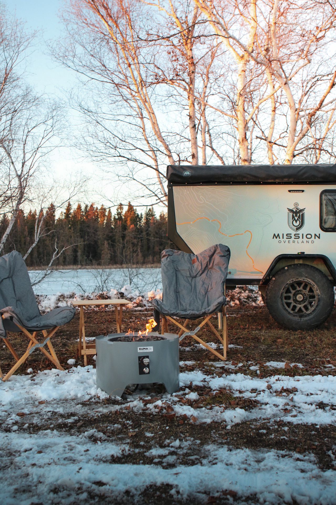 SUV and winter camping setup with tent and chairs in snowy landscape at sunset.