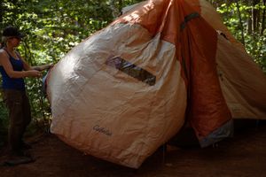 A tent is being set up at a wooded campsite during daytime.