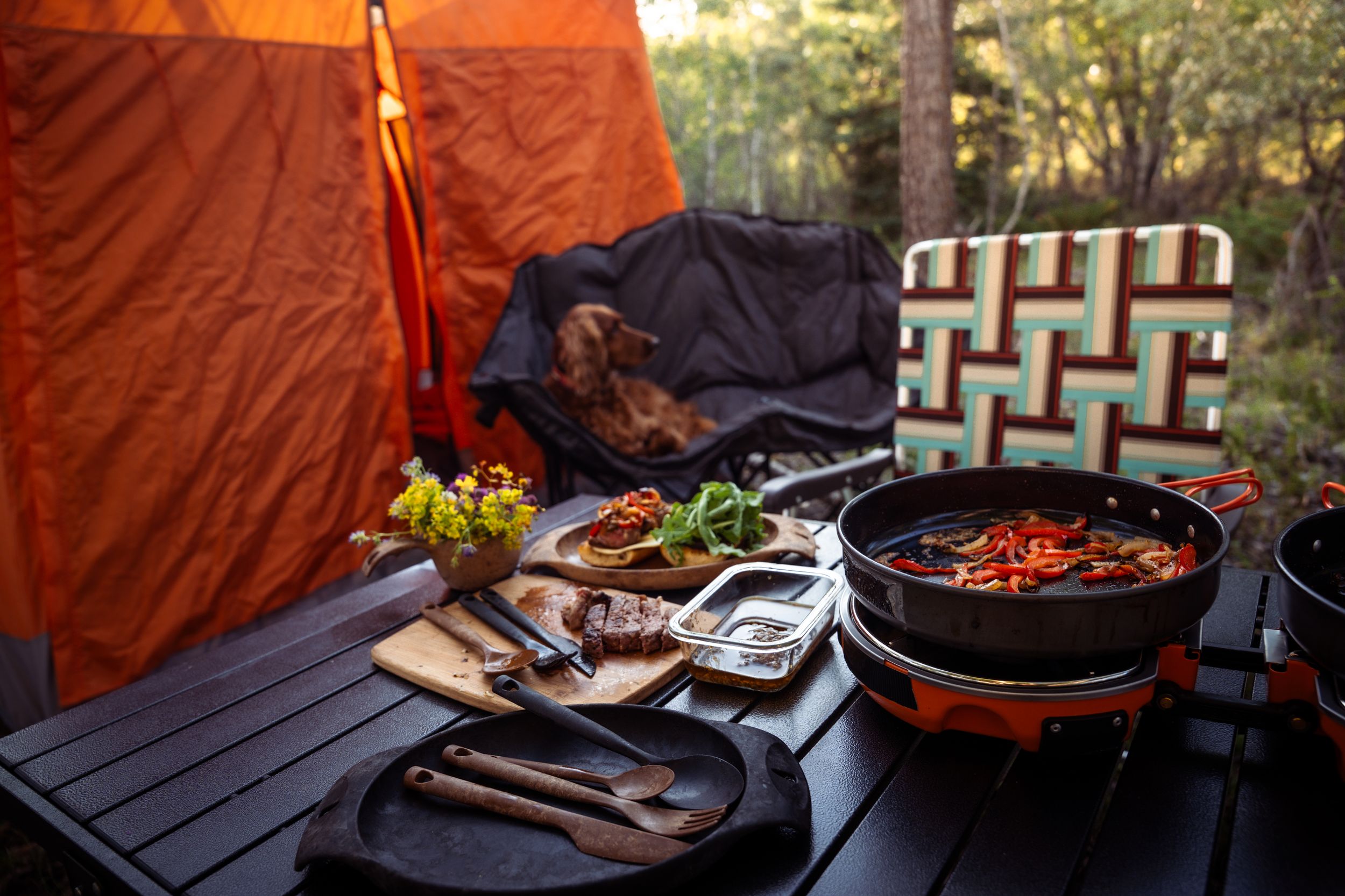 Cast iron pans and grilling accessories arranged on a wooden table during an outdoor cooking session at sunset.