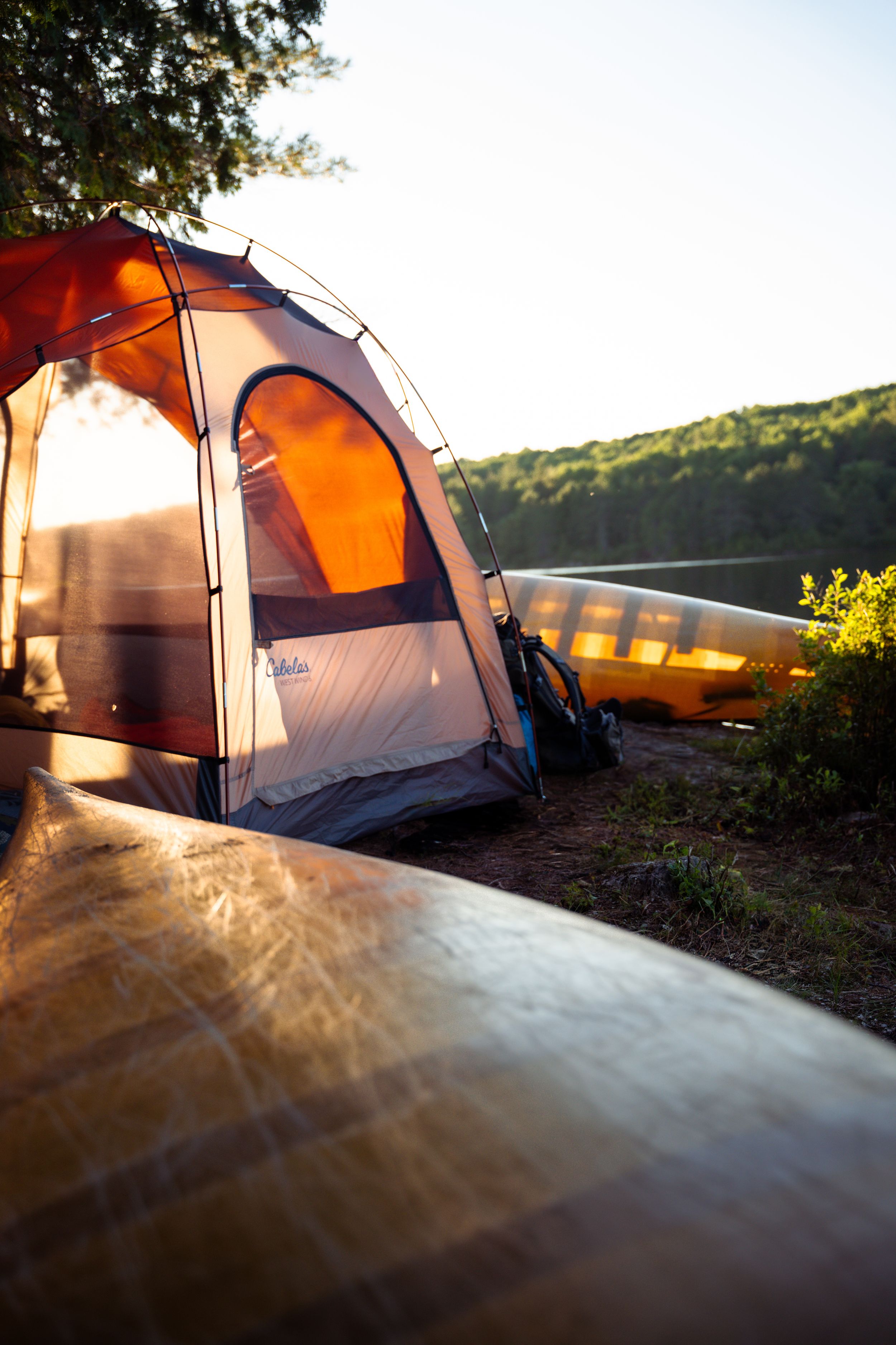 Orange camping tent glows from within at dusk, casting warm light across the surrounding grassy hillside in summer.