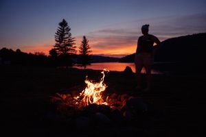 Silhouetted figures gather around a blazing campfire against a vibrant sunset backdrop on the water.