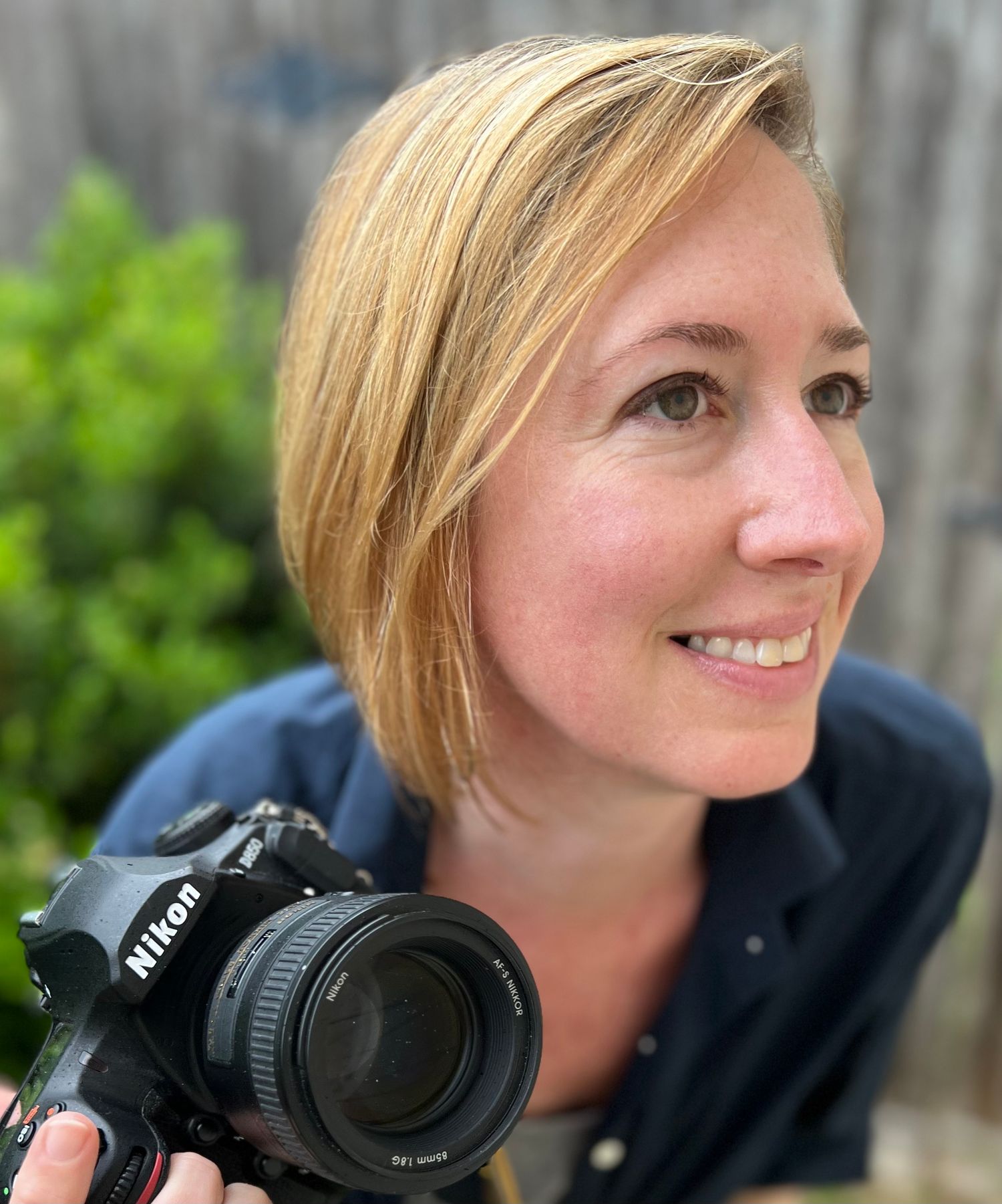 A photographer with blonde hair smiles while holding a Nikon camera against a blurred green outdoor background.