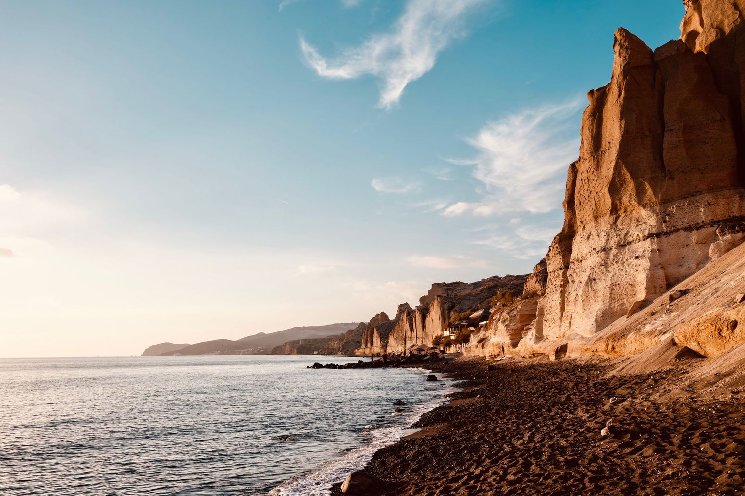 Dramatic coastal landscape with tall rocky cliffs meeting a dark pebble beach and blue ocean.