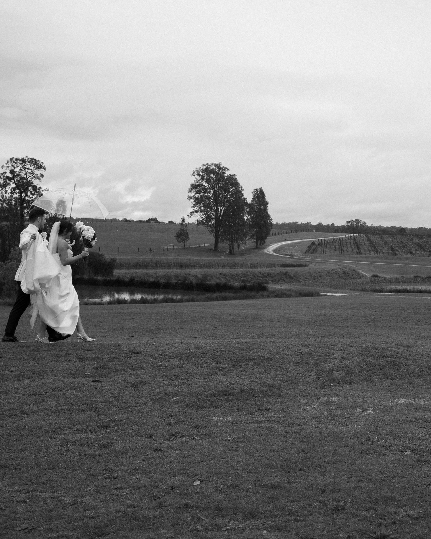 Black and white photo of newlyweds walking across a scenic countryside golf course.