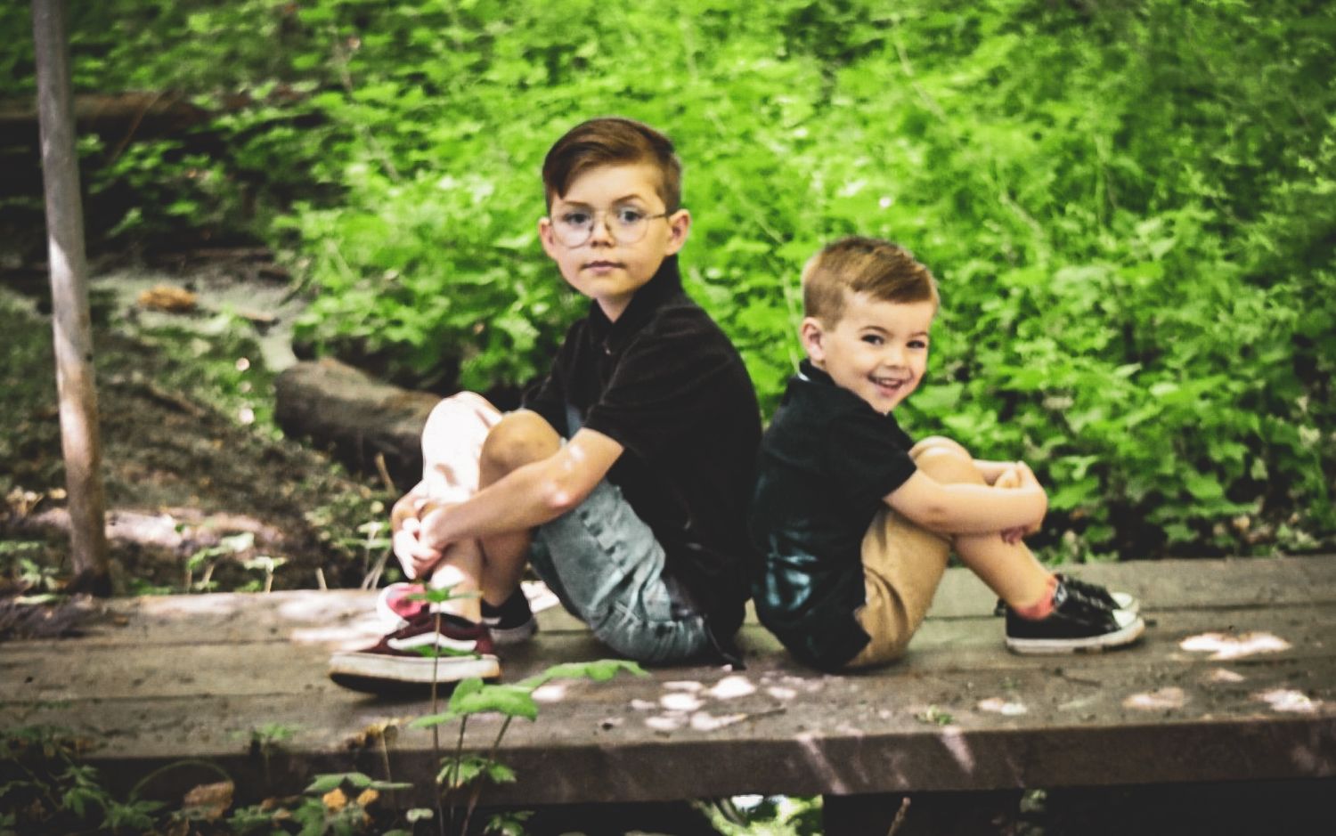 Two boys sitting back-to-back on a log in a wooded area.