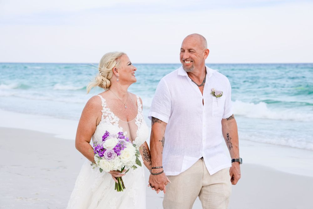 A couple in white beach wedding attire walking along the shoreline with a purple bouquet.