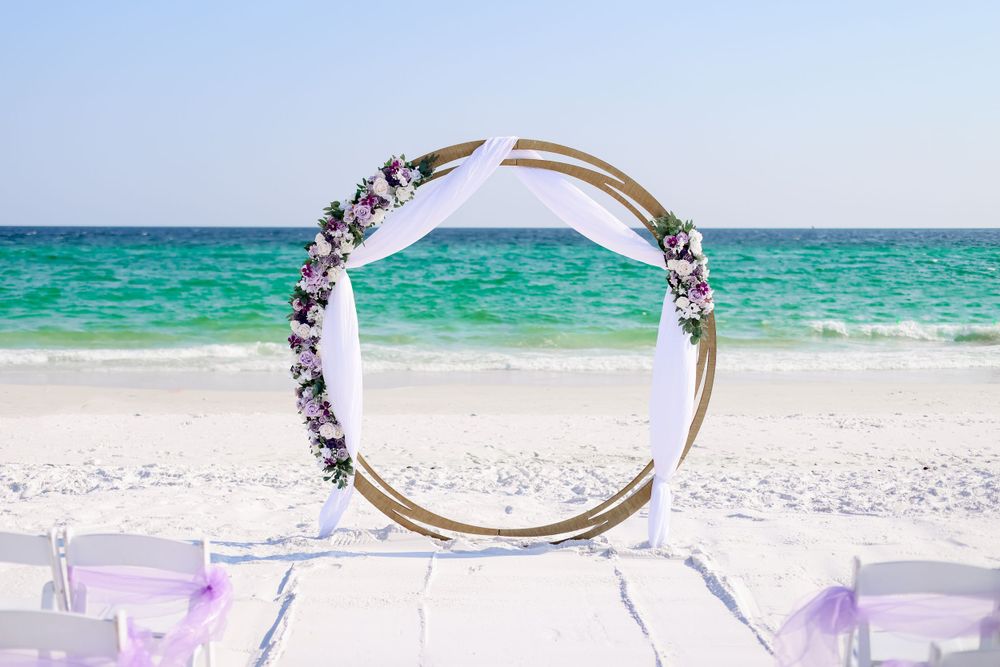 Circular wedding arch decorated with flowers on a beach with turquoise water backdrop.
