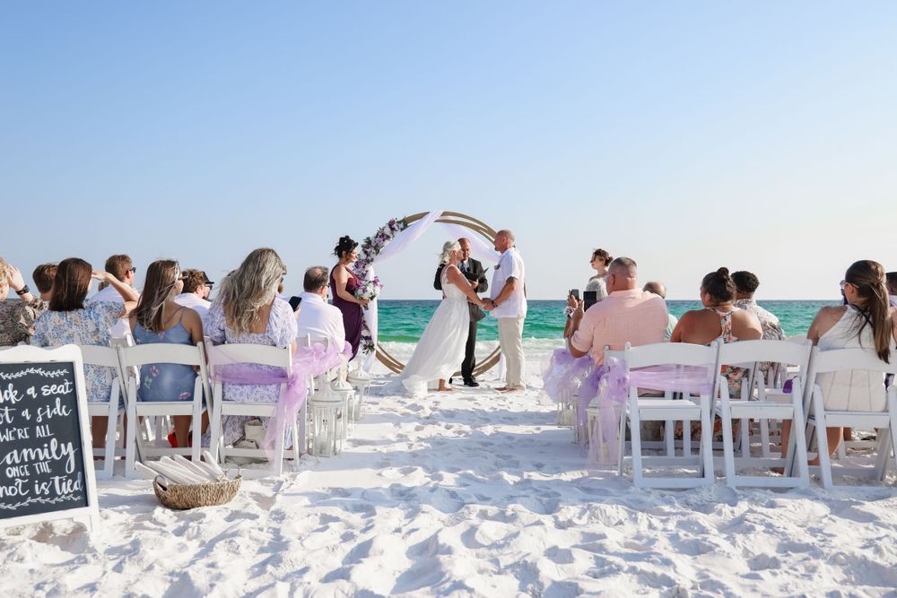 Wedding guests enjoying a beach ceremony with white chairs and floral arch decorations.