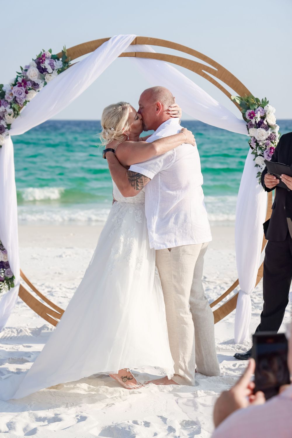 A beach wedding ceremony under a wooden arch decorated with purple and white flowers on a sunny day.