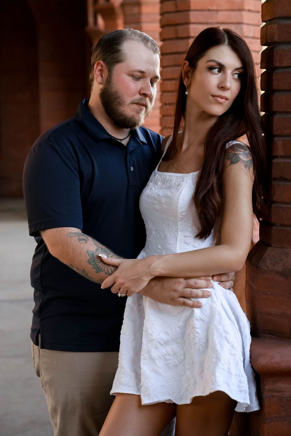 Couple in casual attire poses intimately against brick columns in romantic outdoor portrait session.