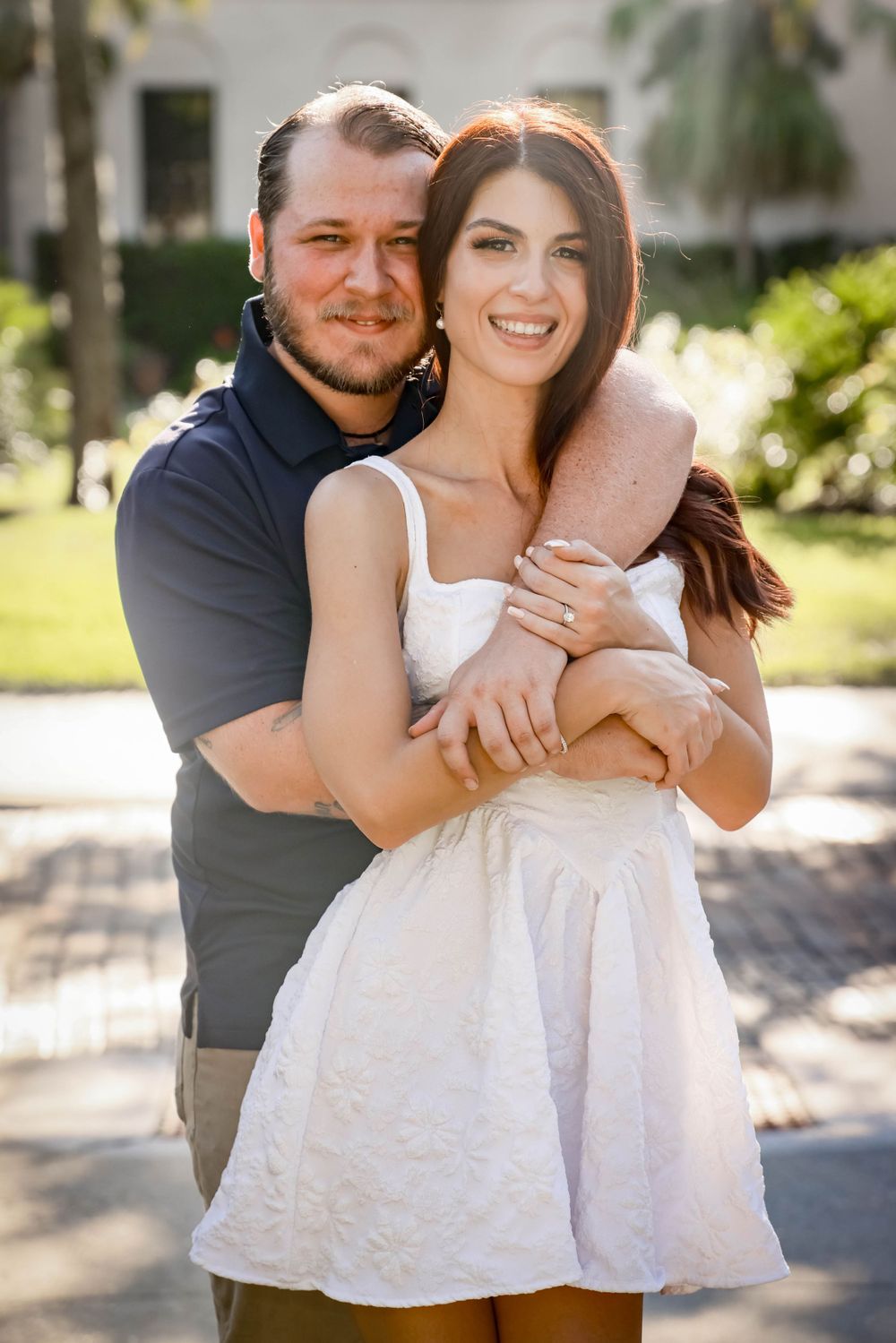 A sunny engagement portrait in front of tropical landscaping and white architecture.