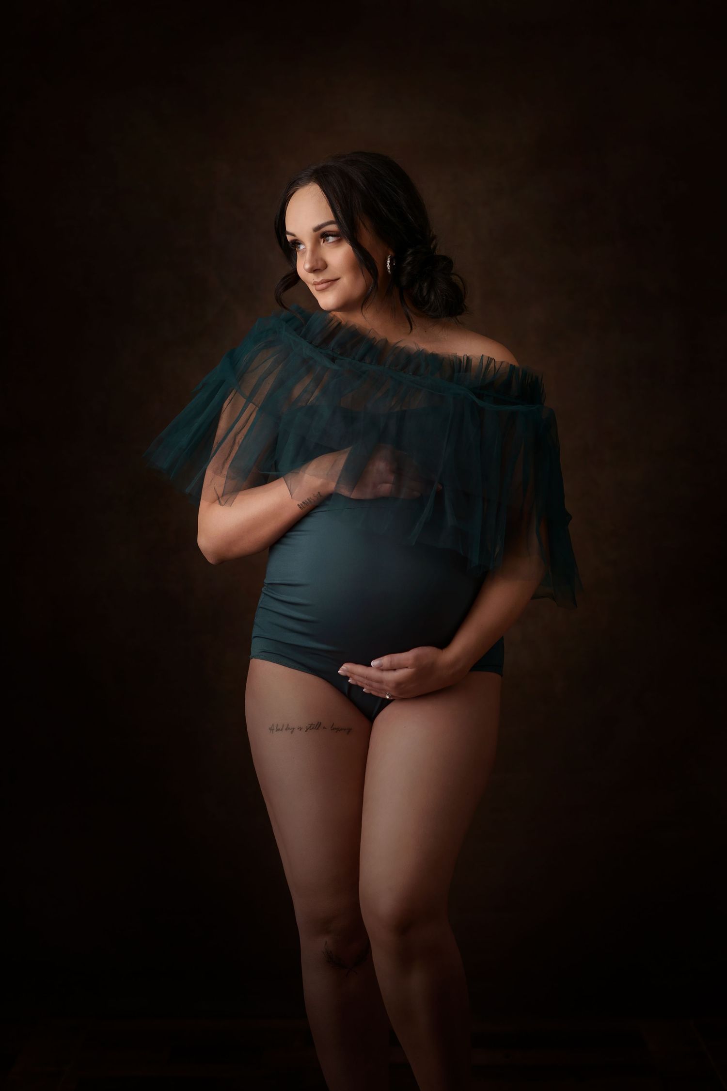 A maternity photo features an elegant teal bodysuit with sheer overlay against a dark dramatic background.  Blackfoot maternity photographer