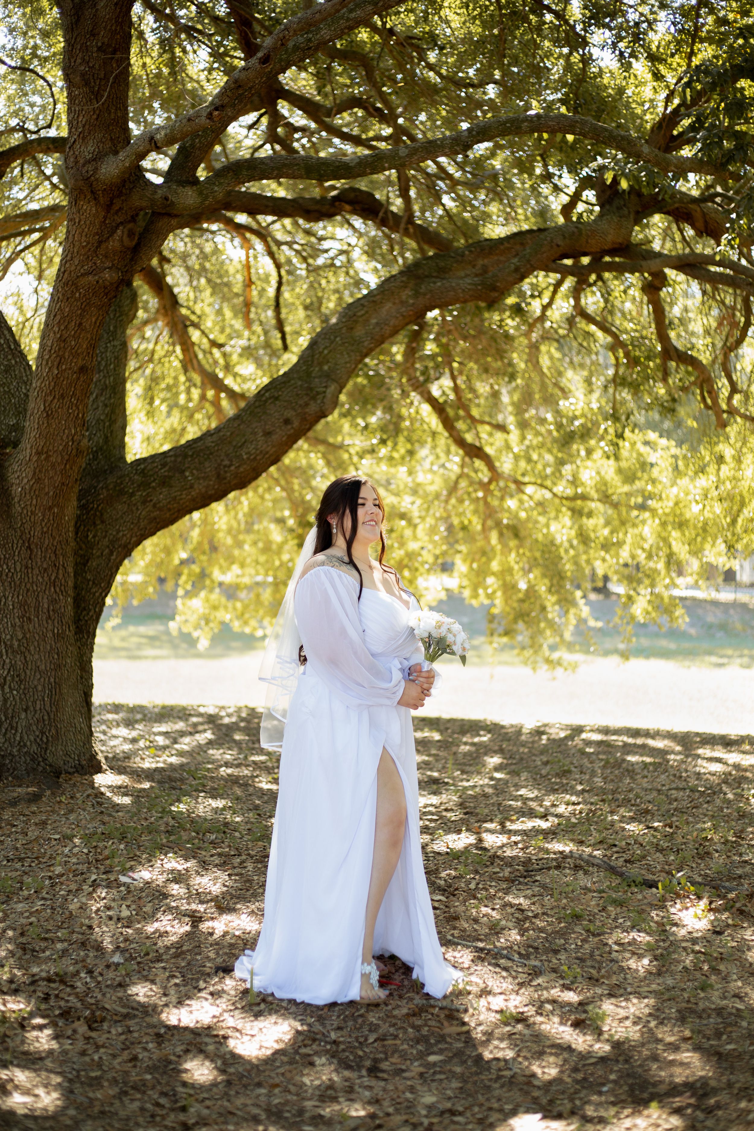 A bride in a flowing white dress stands under a majestic oak tree with golden sunlight filtering through.
