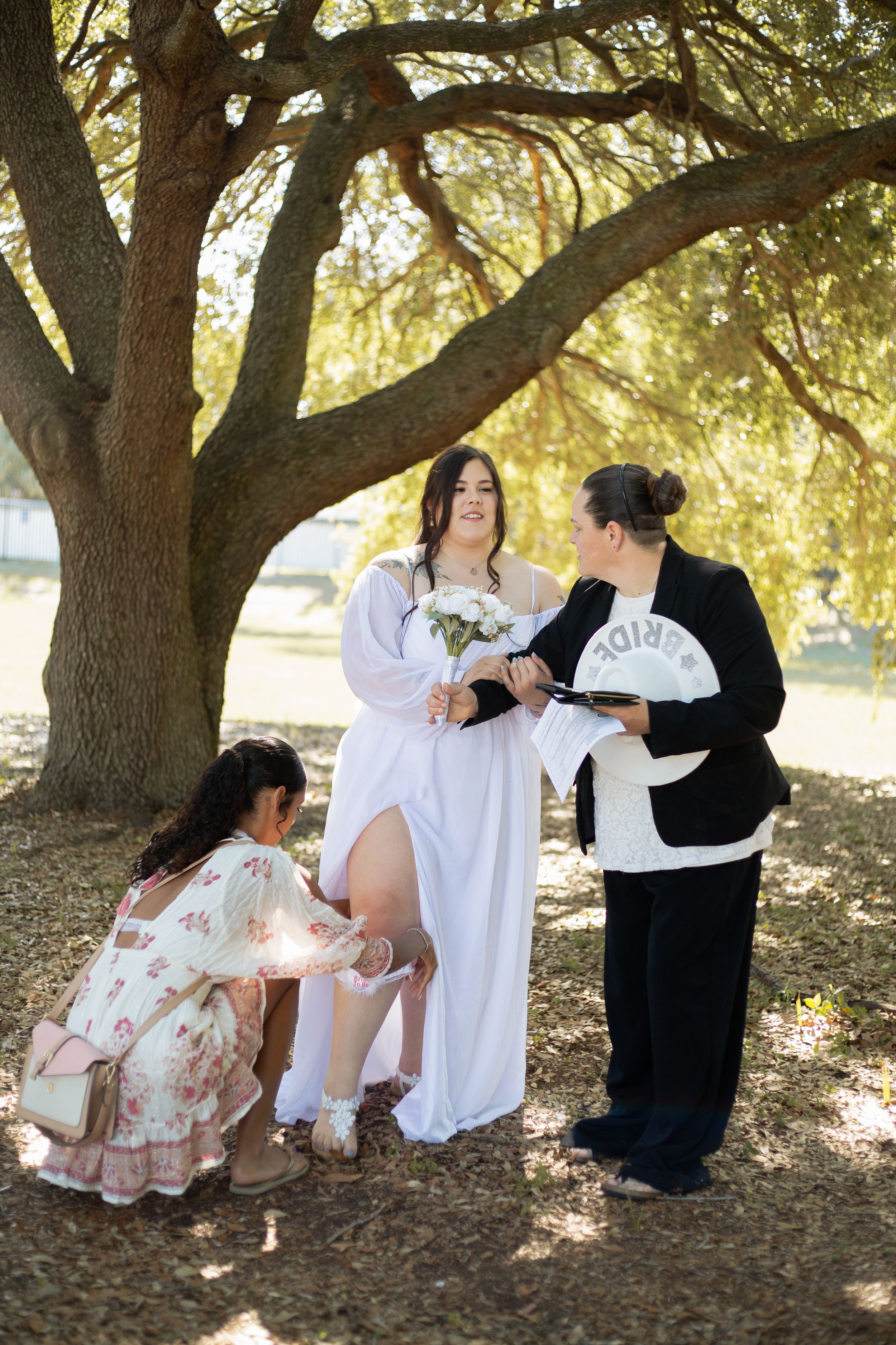 Wedding preparation scene under an oak tree with helpers adjusting the bride's attire.