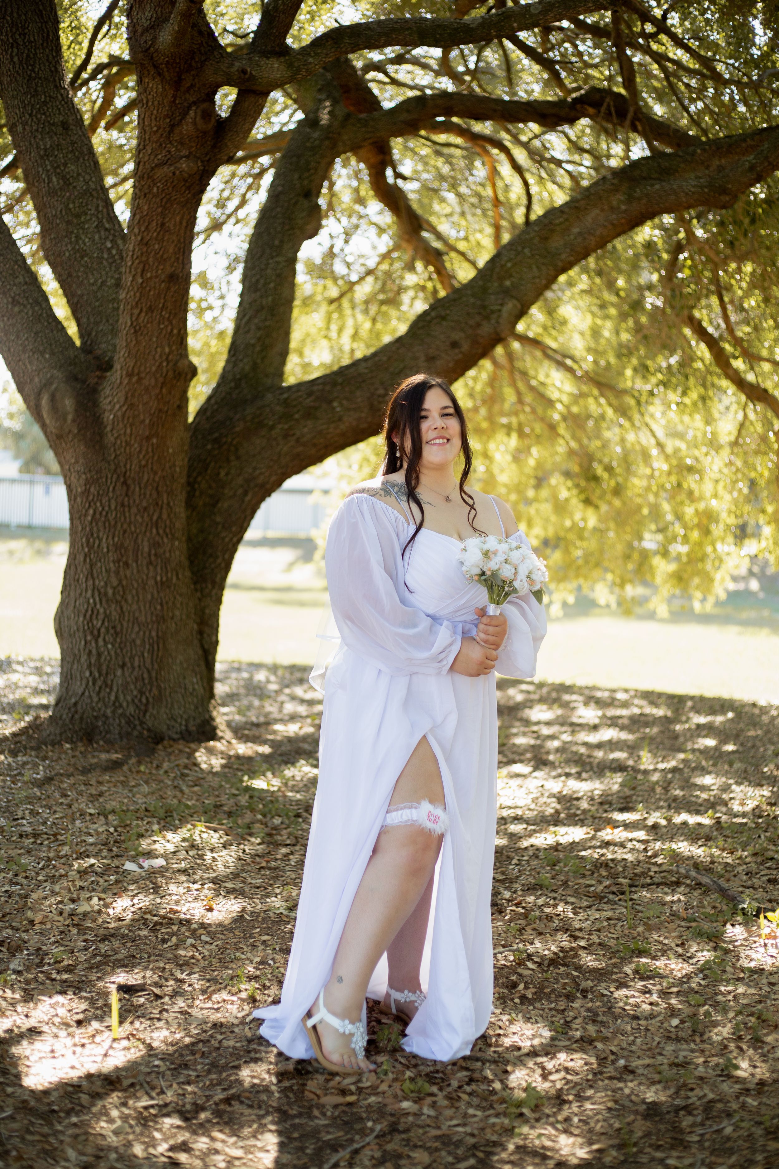 A wedding portrait under a large oak tree featuring an ethereal white off-shoulder dress and bouquet.