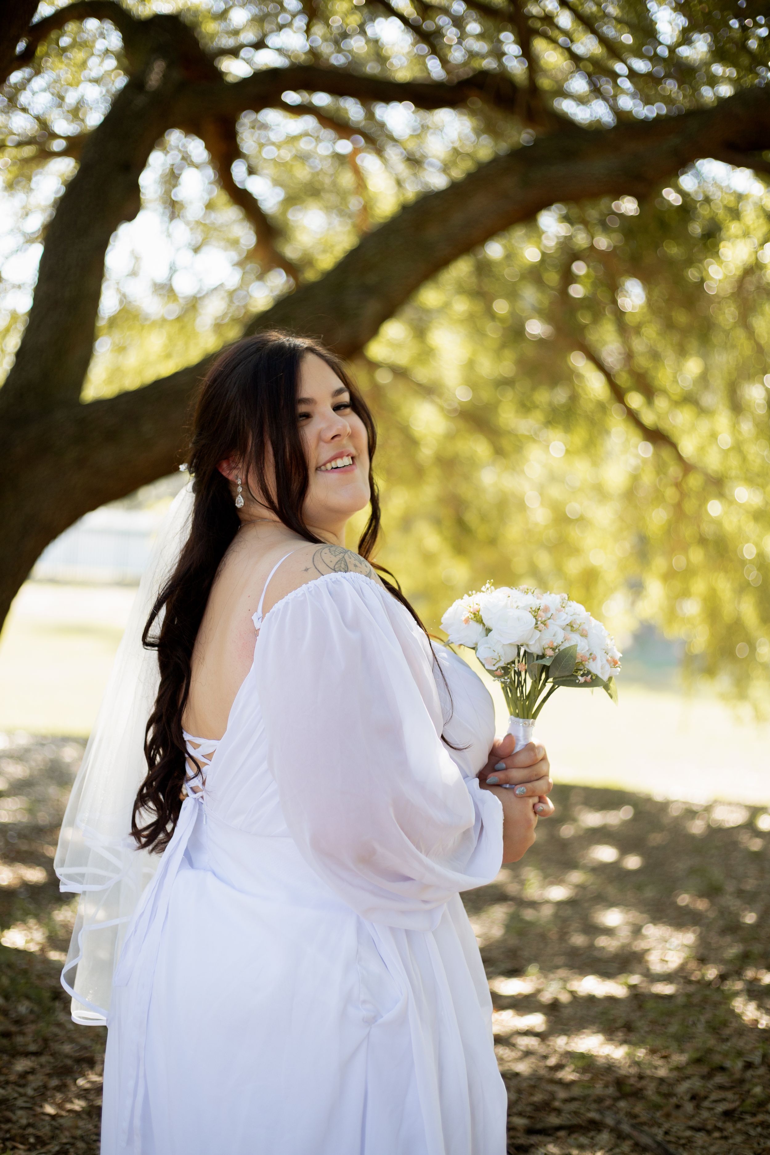 Romantic bridal portrait with flowing white dress and white bouquet under a sun-dappled tree.