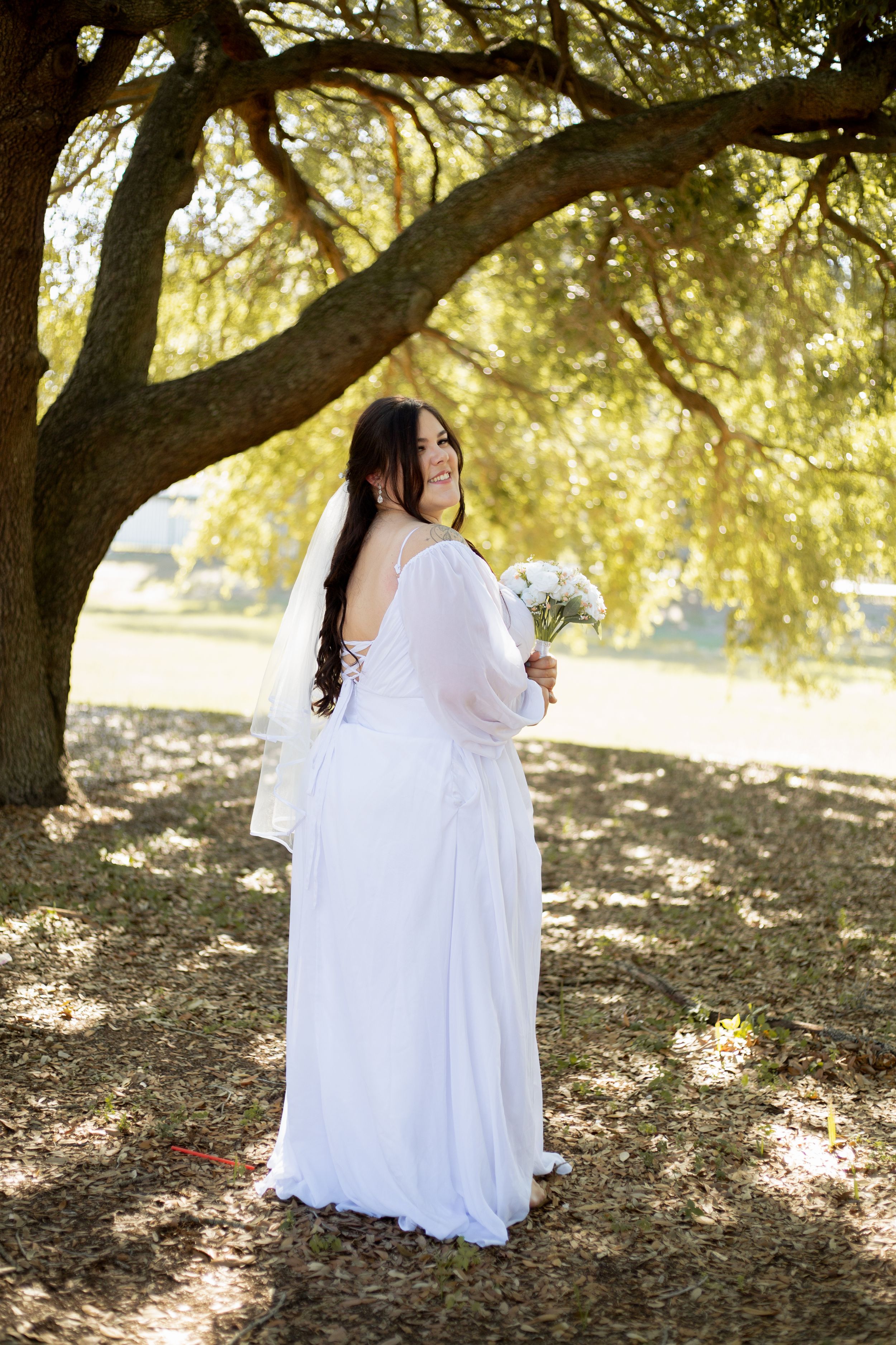 Full-length bridal portrait showcasing a flowing white gown and bouquet in dappled sunlight.