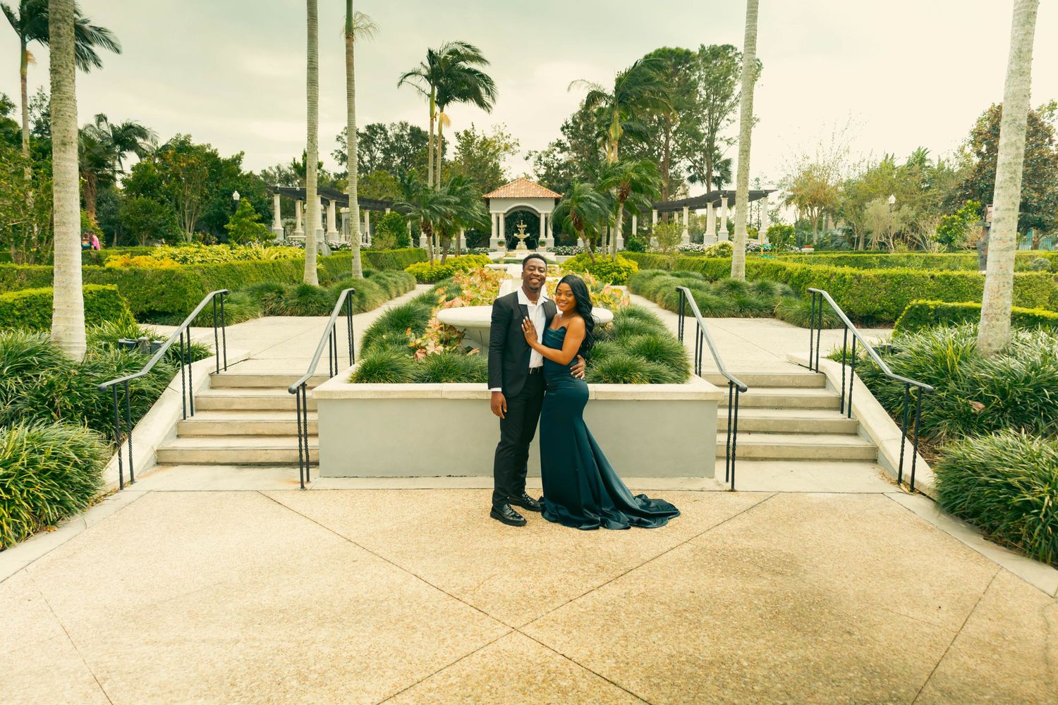 Elegant couple posing in formal attire before a grand estate garden with palm trees and manicured hedges.
