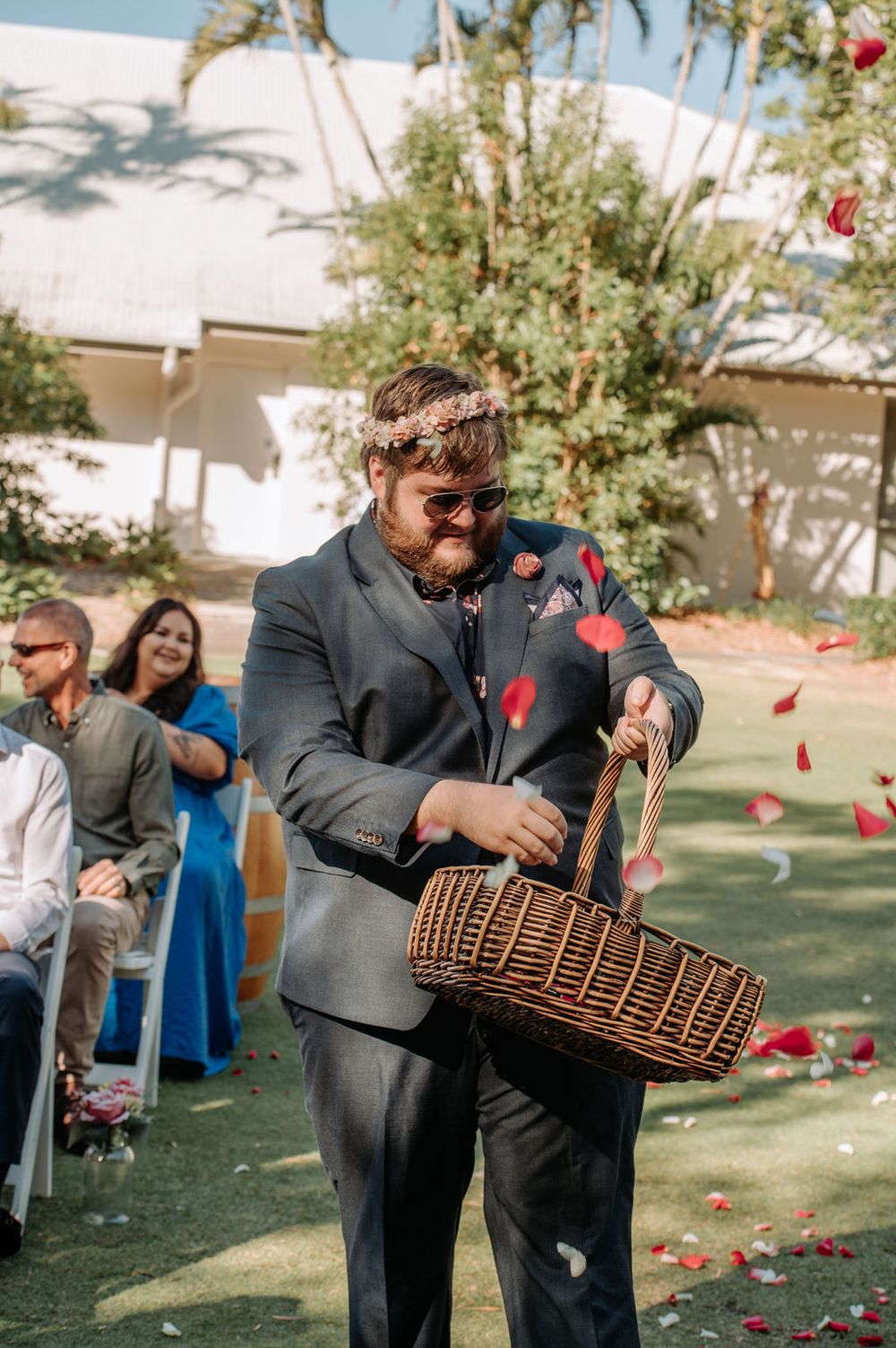Wedding guest in a grey suit scatters rose petals from a wicker basket during outdoor ceremony.