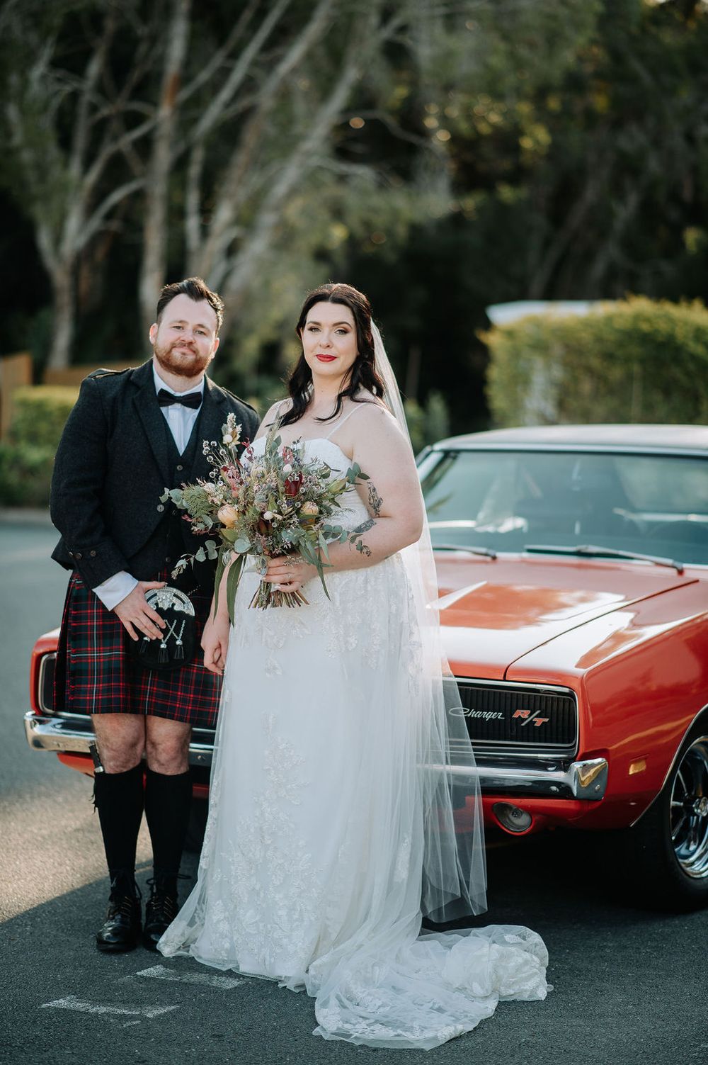 A wedding couple poses with a classic red car as the groom wears a traditional Scottish kilt and the bride holds a bouquet.