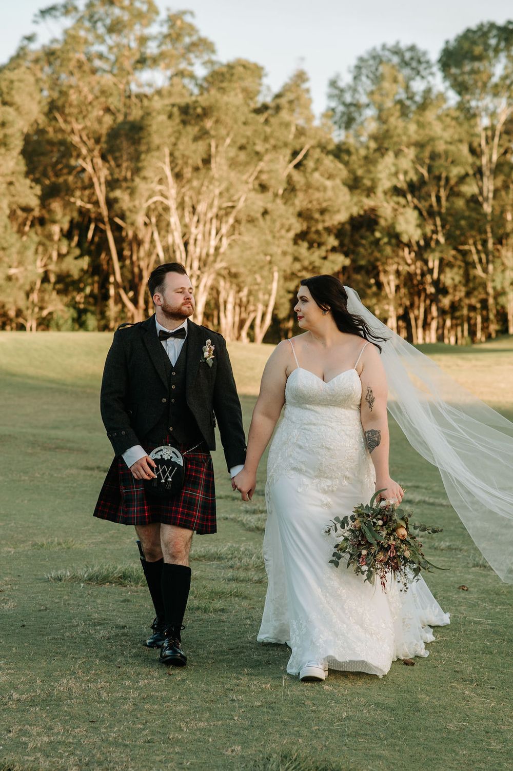 A couple walks hand in hand on a grassy field with the groom in a kilt and bride in a flowing white dress.