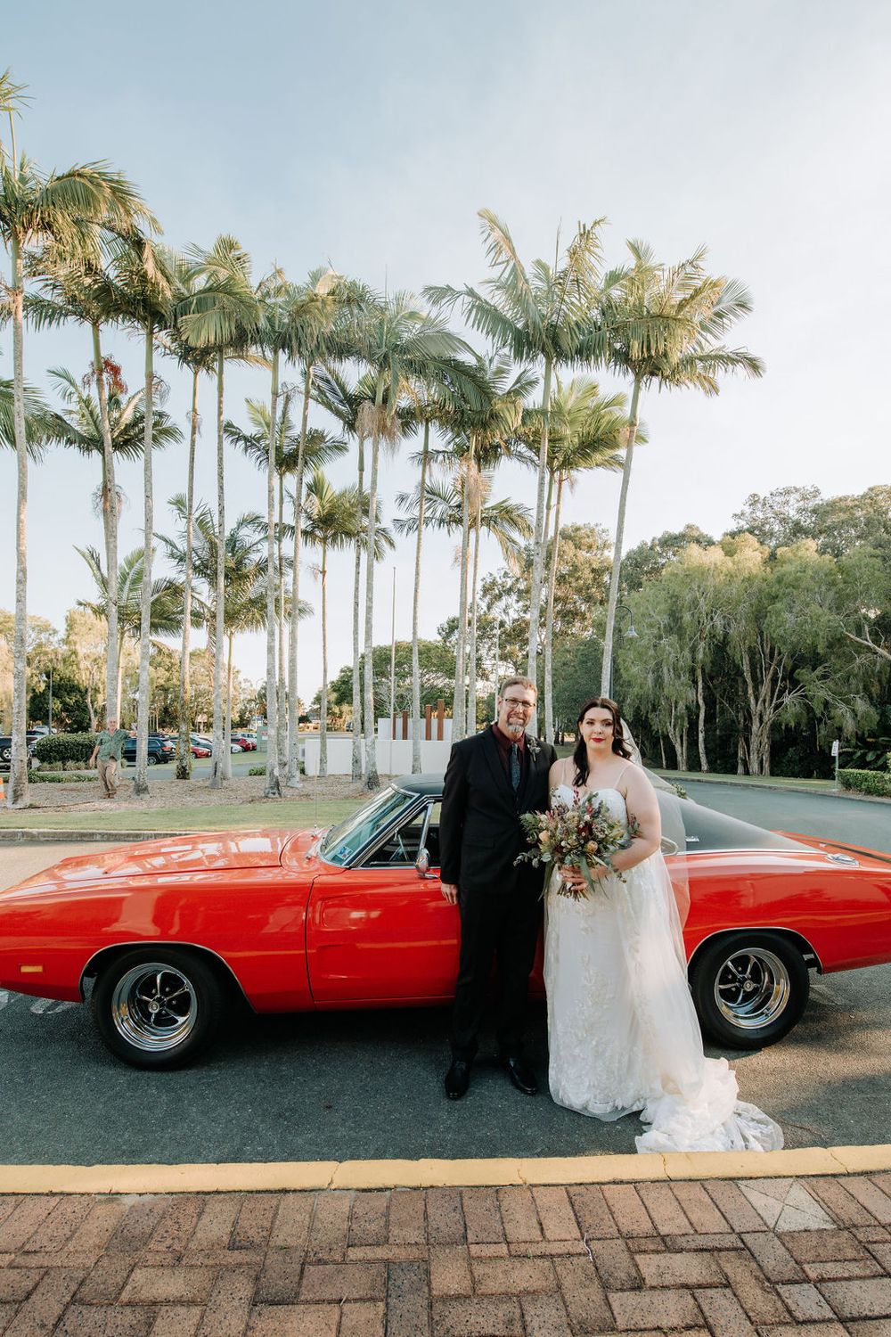 Wedding couple poses with classic red Dodge Charger against tropical palm tree backdrop.
