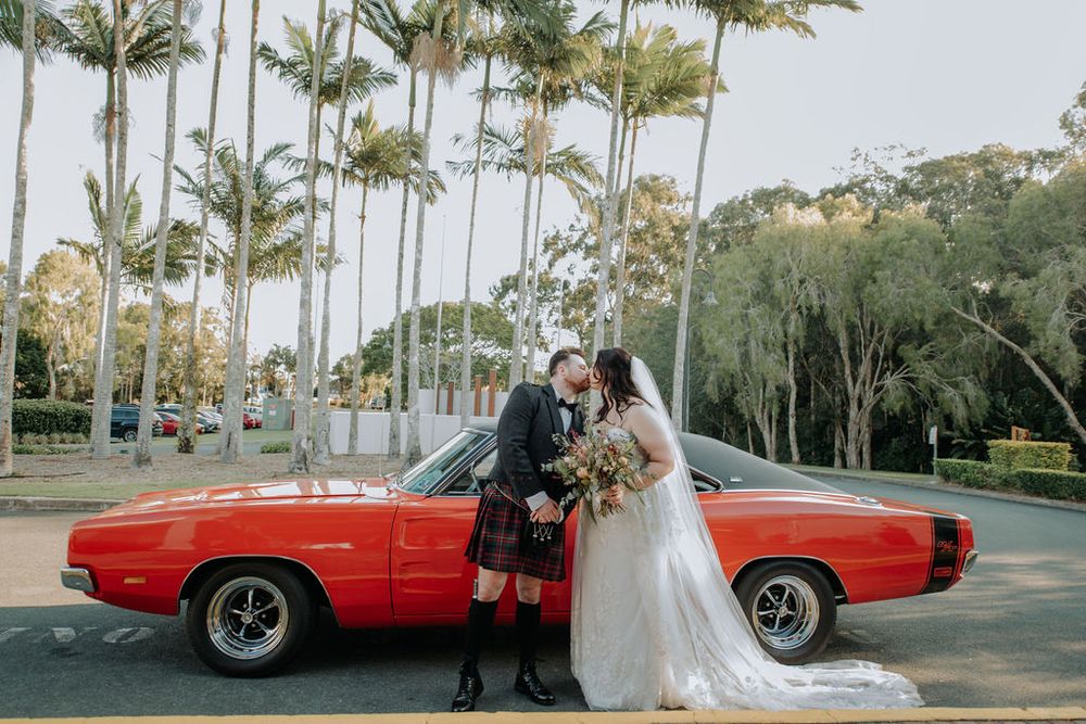 Newlyweds pose with vintage red muscle car against backdrop of palm trees.