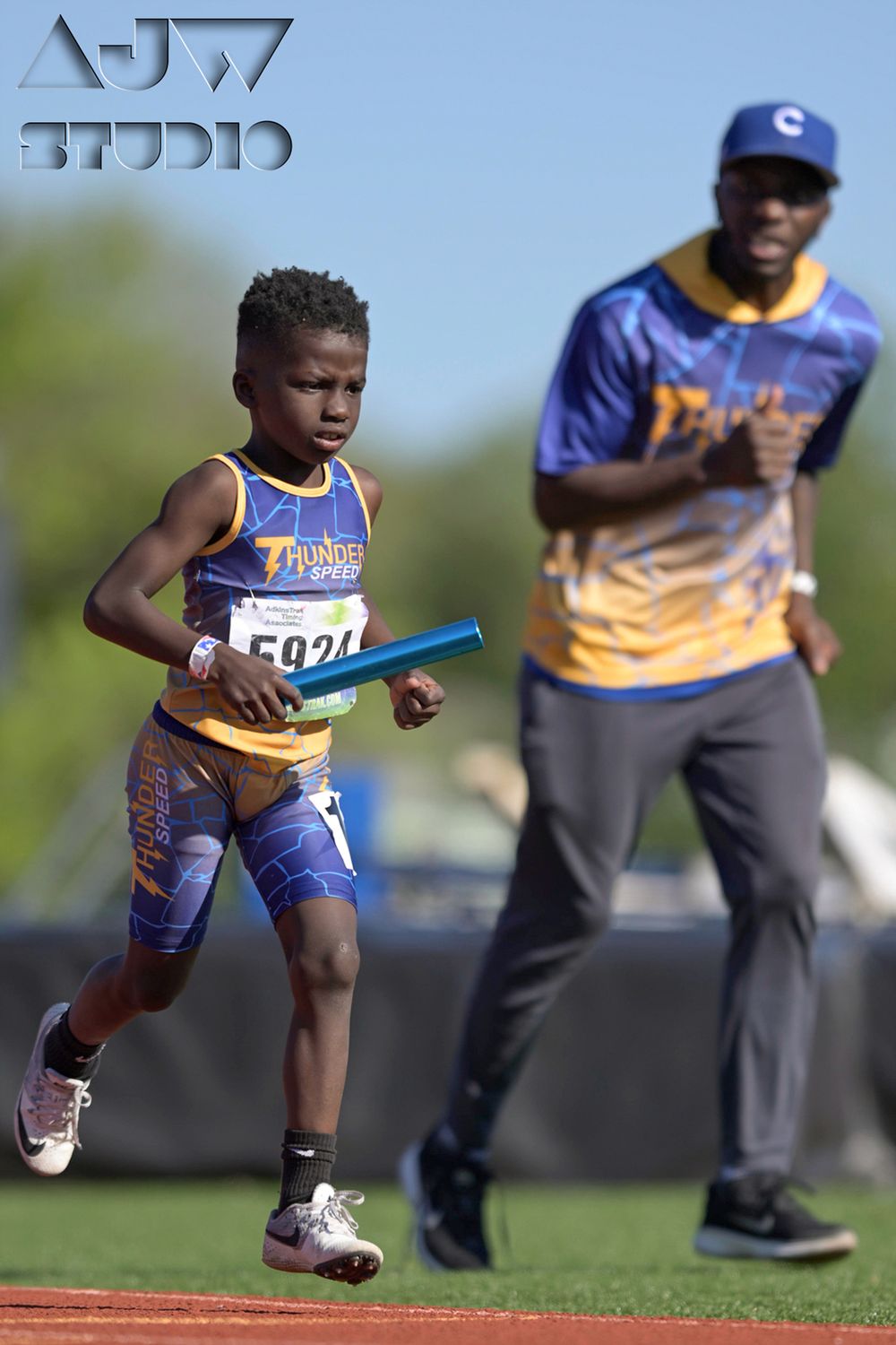 Young athlete (shadowed by his coach) runs with relay baton during track meet competition.