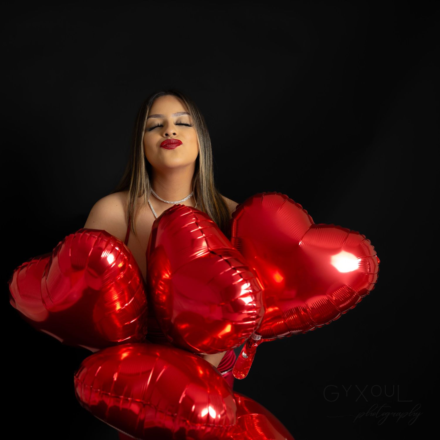 Studio portrait with cluster of red metallic heart balloons against black background.