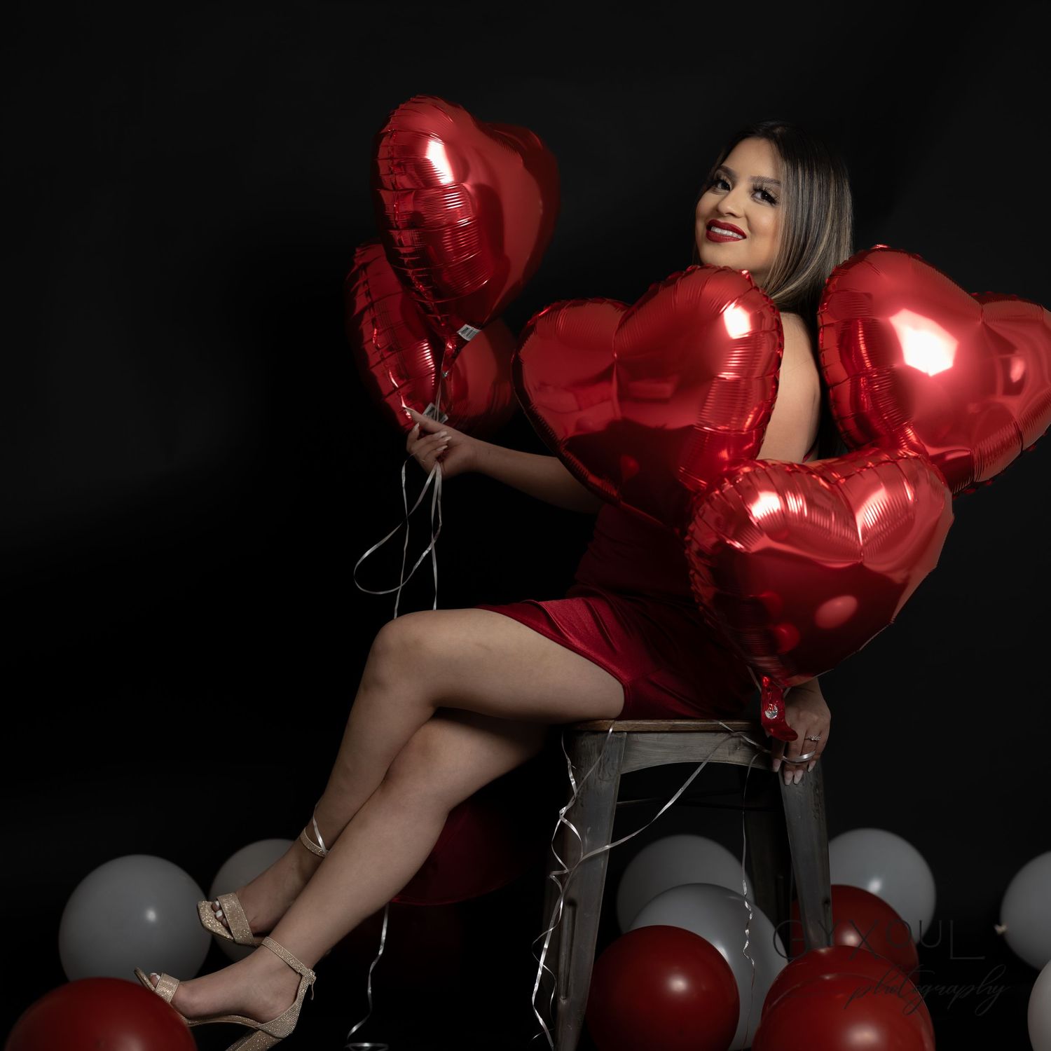 Glamorous Valentine's themed photo with red dress and heart balloons on chair.