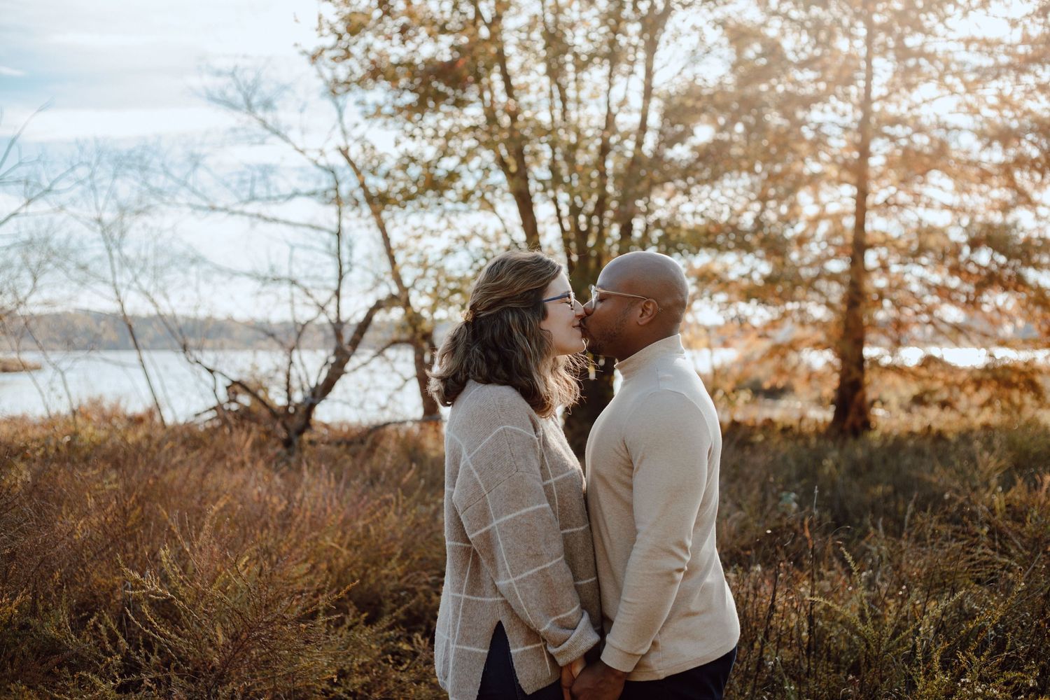 A couple embraces tenderly against an autumn landscape backdrop near water.
