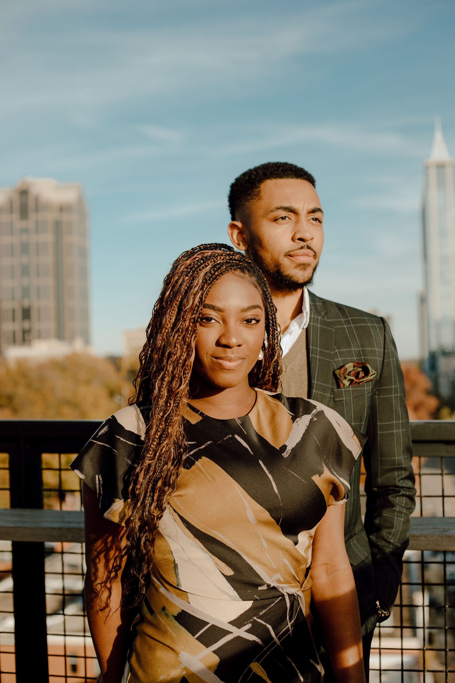 A stylishly dressed couple poses together on a rooftop terrace during golden hour with city buildings in the background.