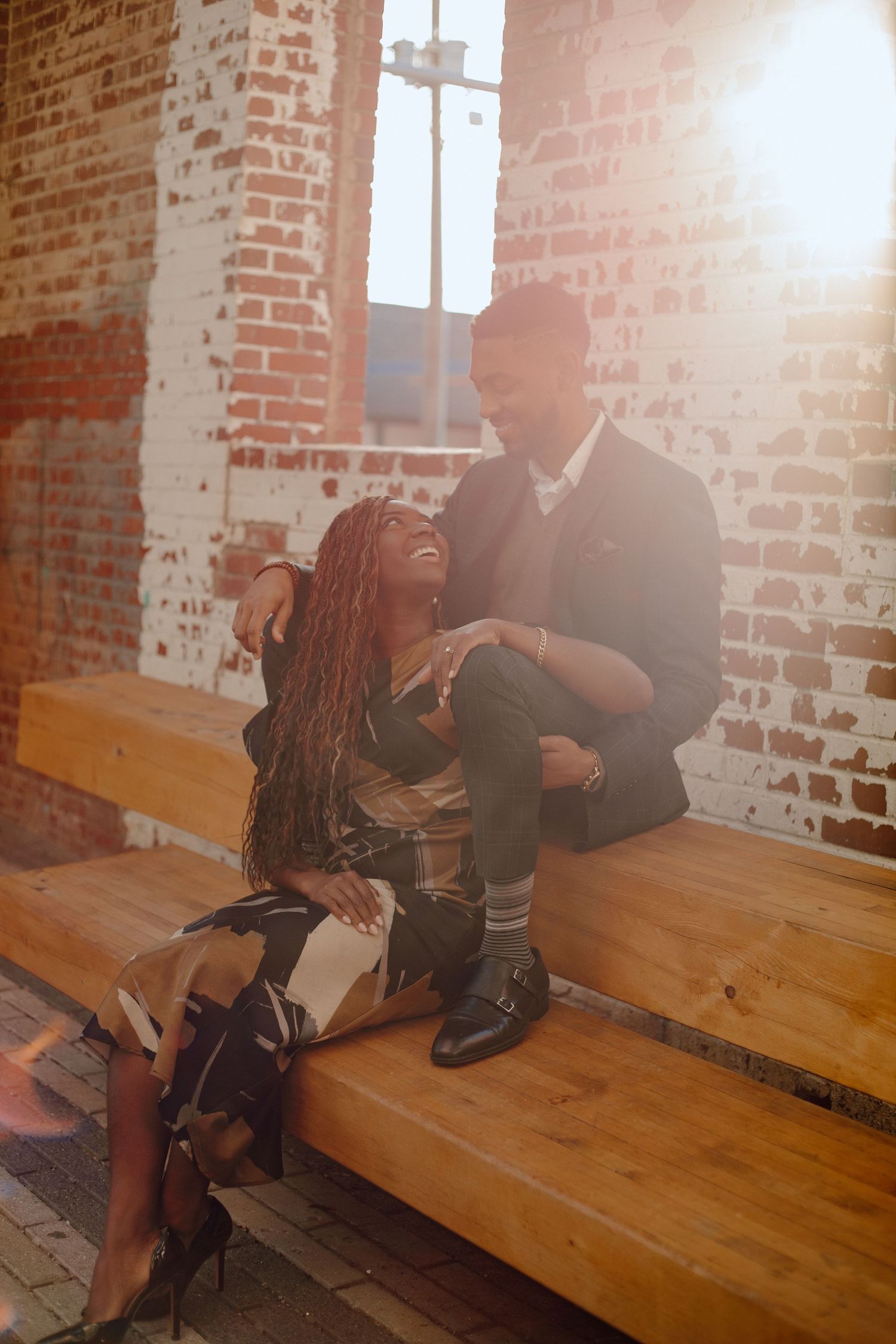 A couple sits together on wooden benches inside a rustic brick building with sunlight streaming through the window.