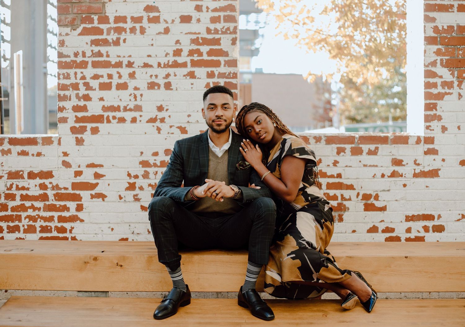 A couple sits close together on a wooden bench against an exposed brick wall during a warm sunset.