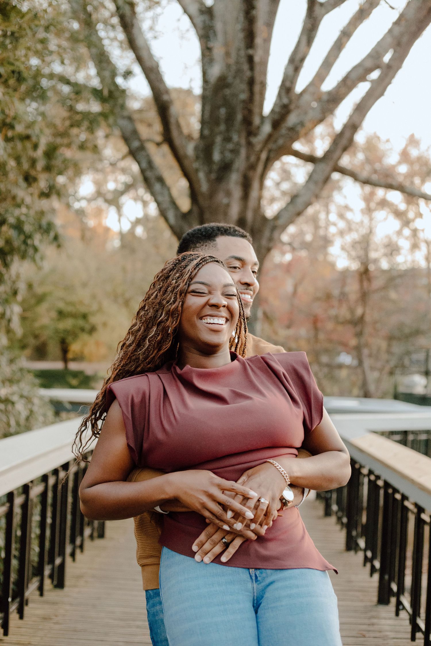 A person in a burgundy top and jeans stands laughing on a wooden bridge surrounded by autumn trees.