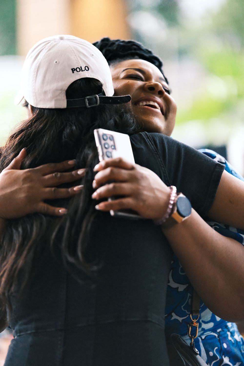 Two friends share an emotional embrace while one wears a white Polo cap at an outdoor gathering.
