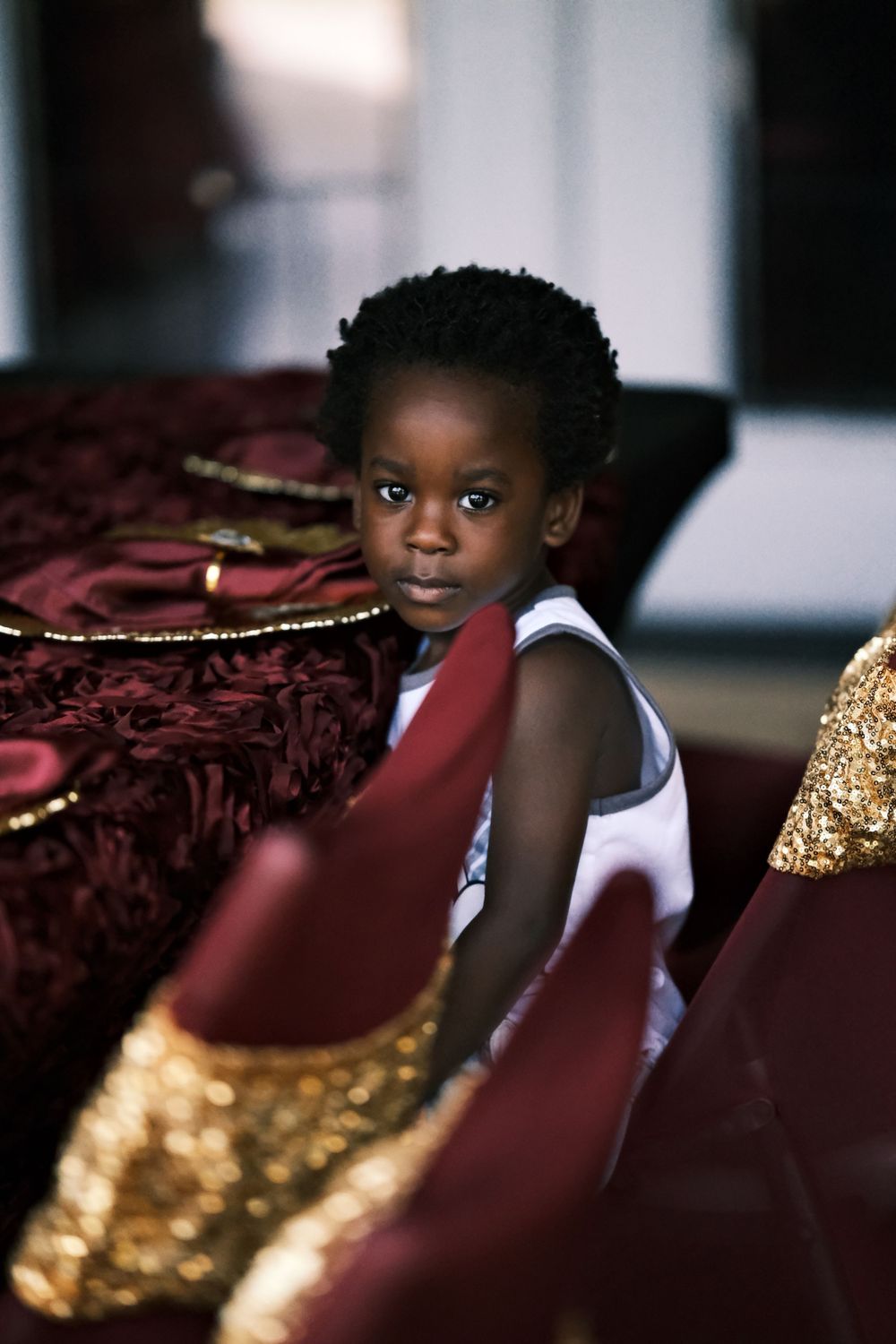 Small child in white tank top poses against luxurious burgundy and gold decor.