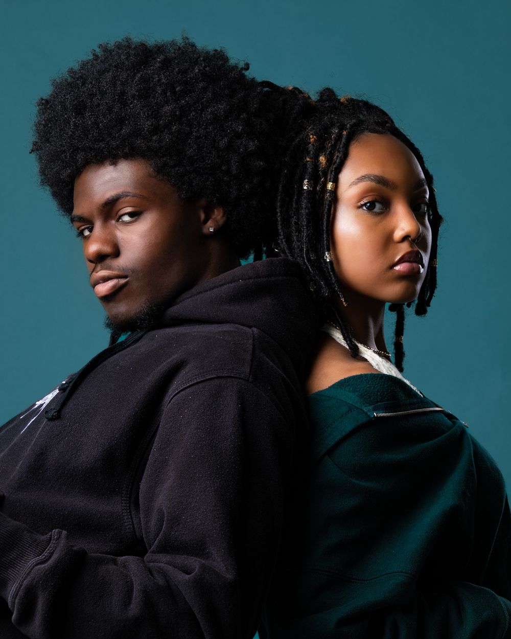 Dramatic studio portrait of two people back-to-back against teal background.
