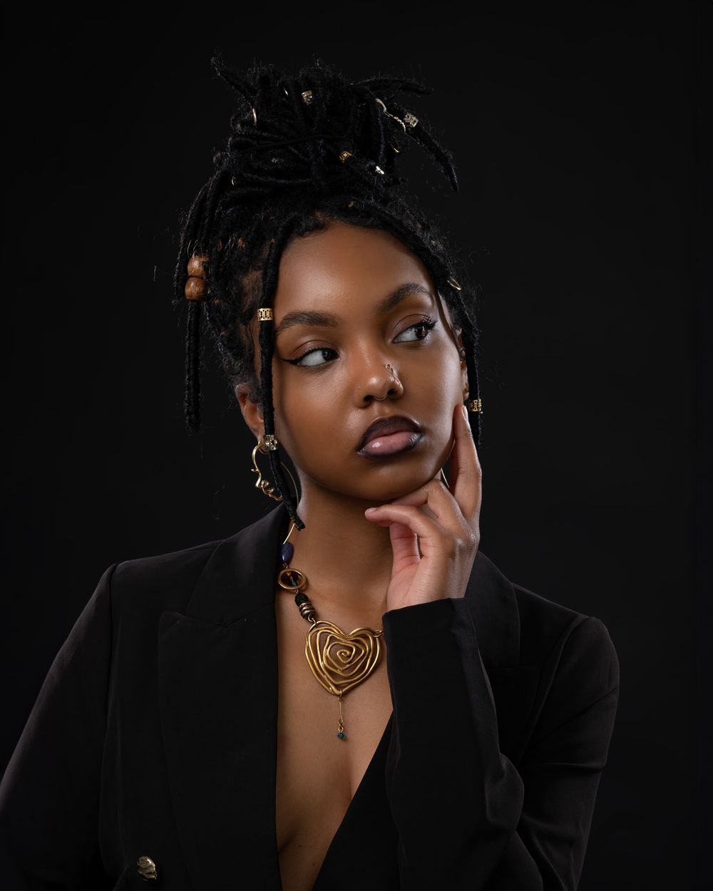 Elegant studio portrait with braided hair styling and gold heart necklace against dark background.