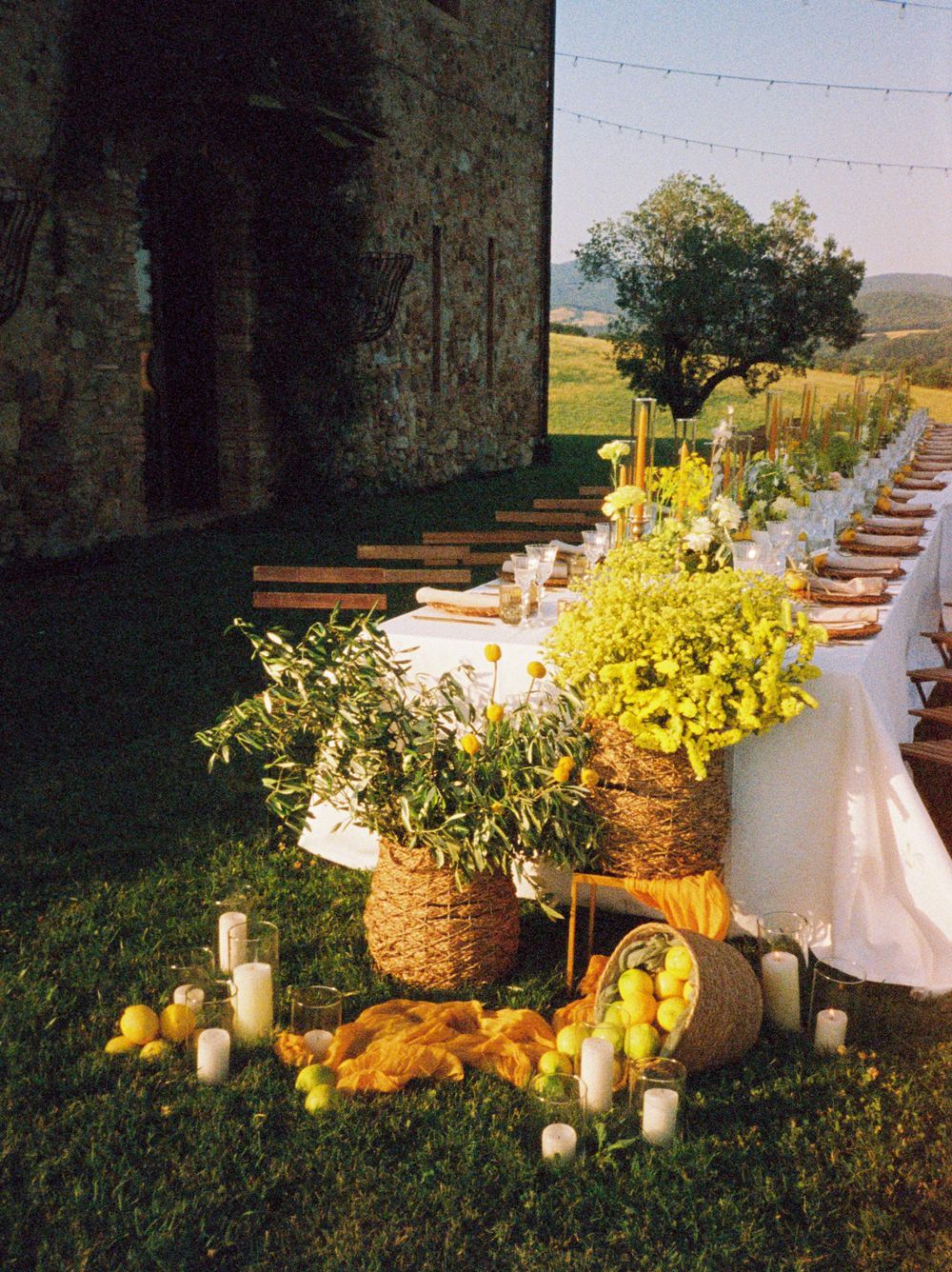 Long outdoor dining table decorated with candles, yellow flowers, and lanterns against a garden backdrop at sunset.