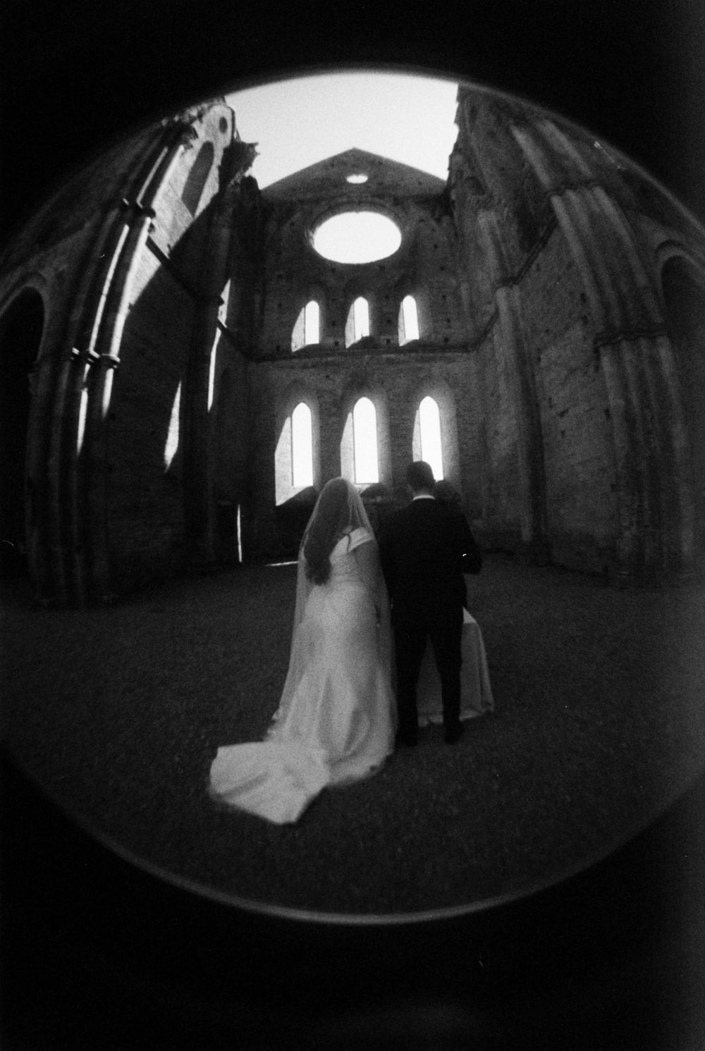A couple in wedding attire stands in silhouette inside a dramatic cathedral with arched windows and natural light.