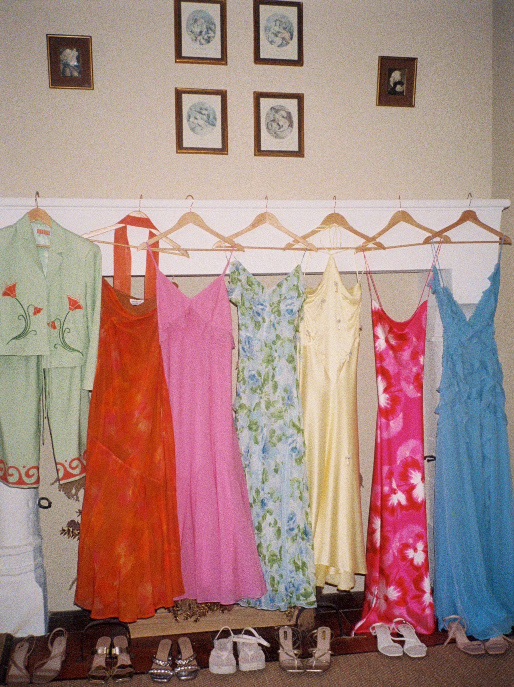 Colorful vintage dresses and a suit hanging on wooden hangers against a beige wall with framed artwork above.