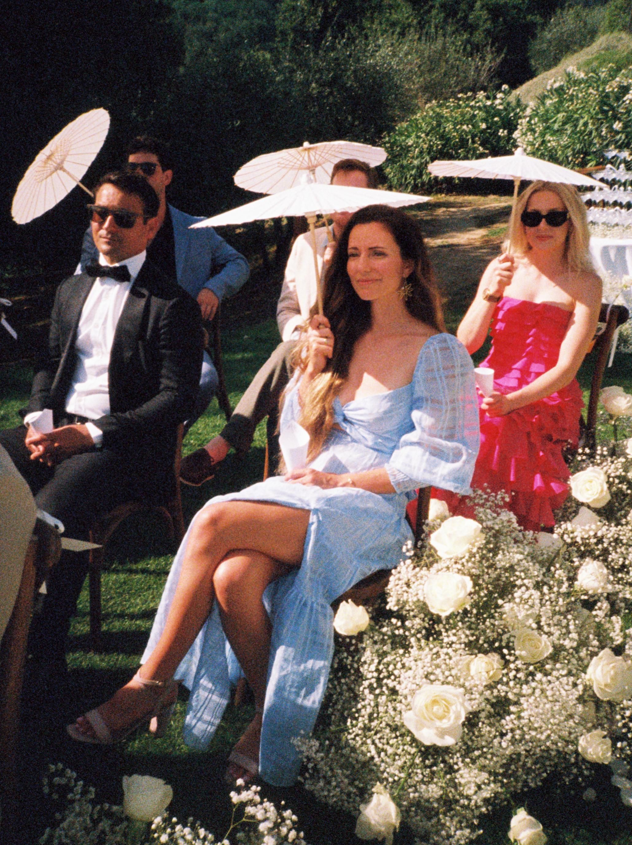 Wedding guests relax outdoors on white chairs with parasols providing shade during a sunny garden ceremony.