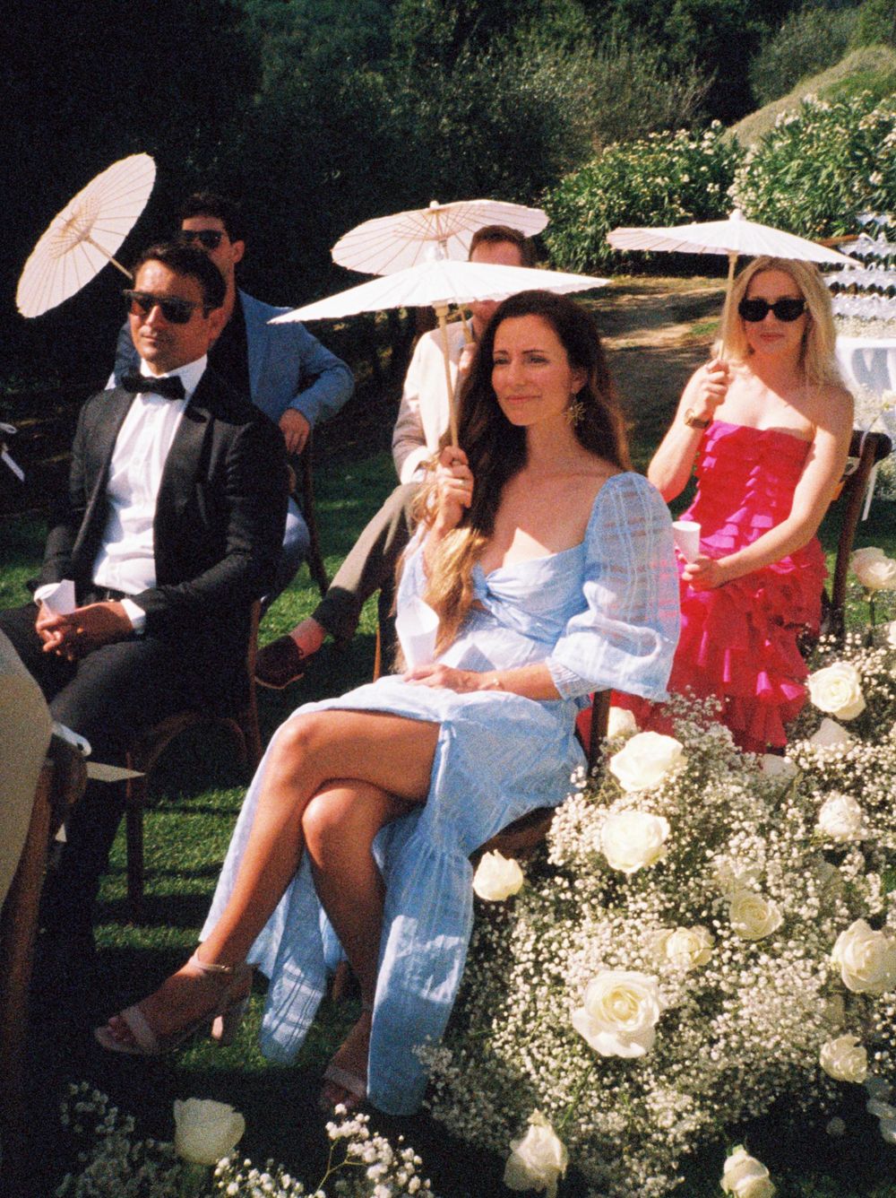 Wedding guests relax outdoors on white chairs with parasols providing shade during a sunny garden ceremony.