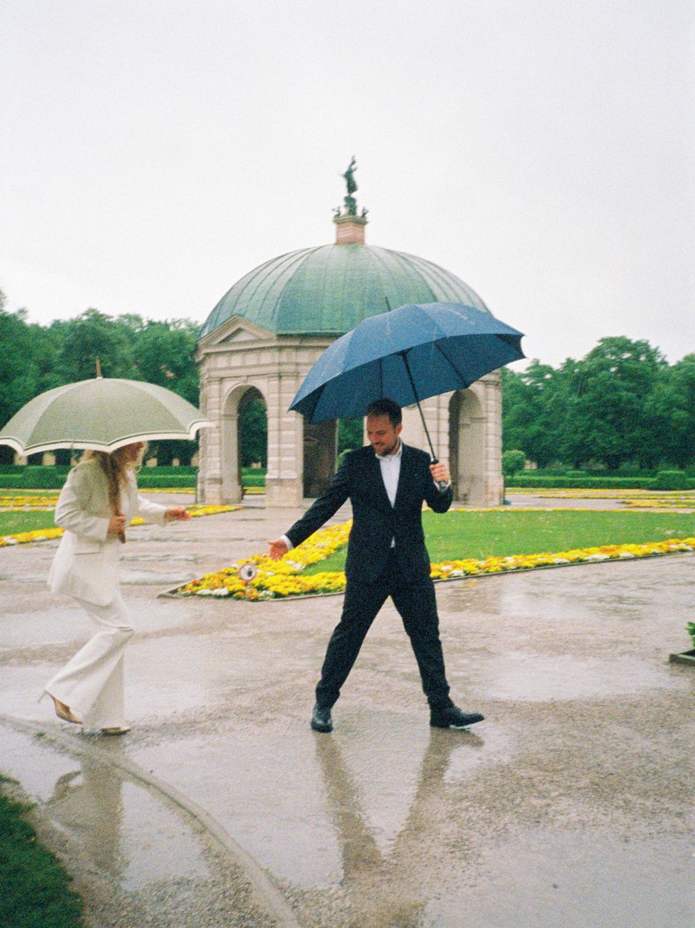 A couple dancing playfully in the rain with umbrellas at an ornate domed structure on a wet stone courtyard.